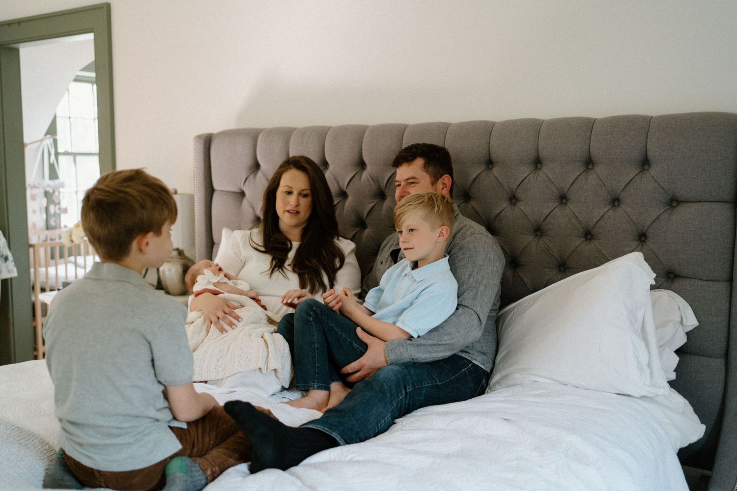A family sits together on a gray tufted headboard bed, sharing a relaxed moment at home.