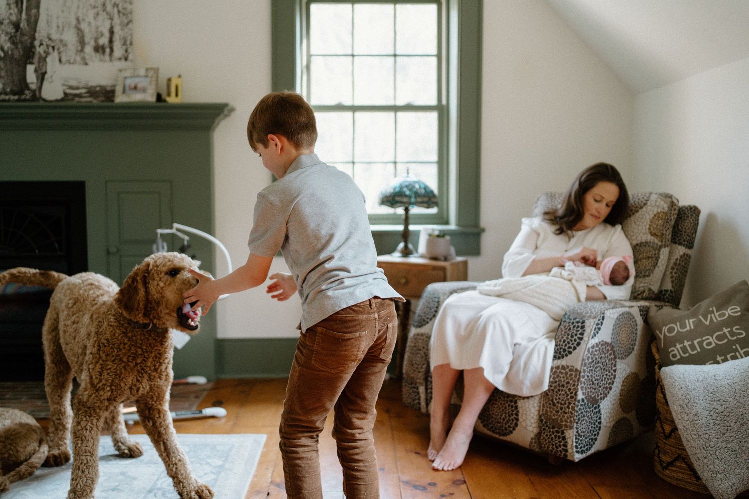 A child plays with a goldendoodle dog while someone rests with an infant in a cozy living room with green walls.
