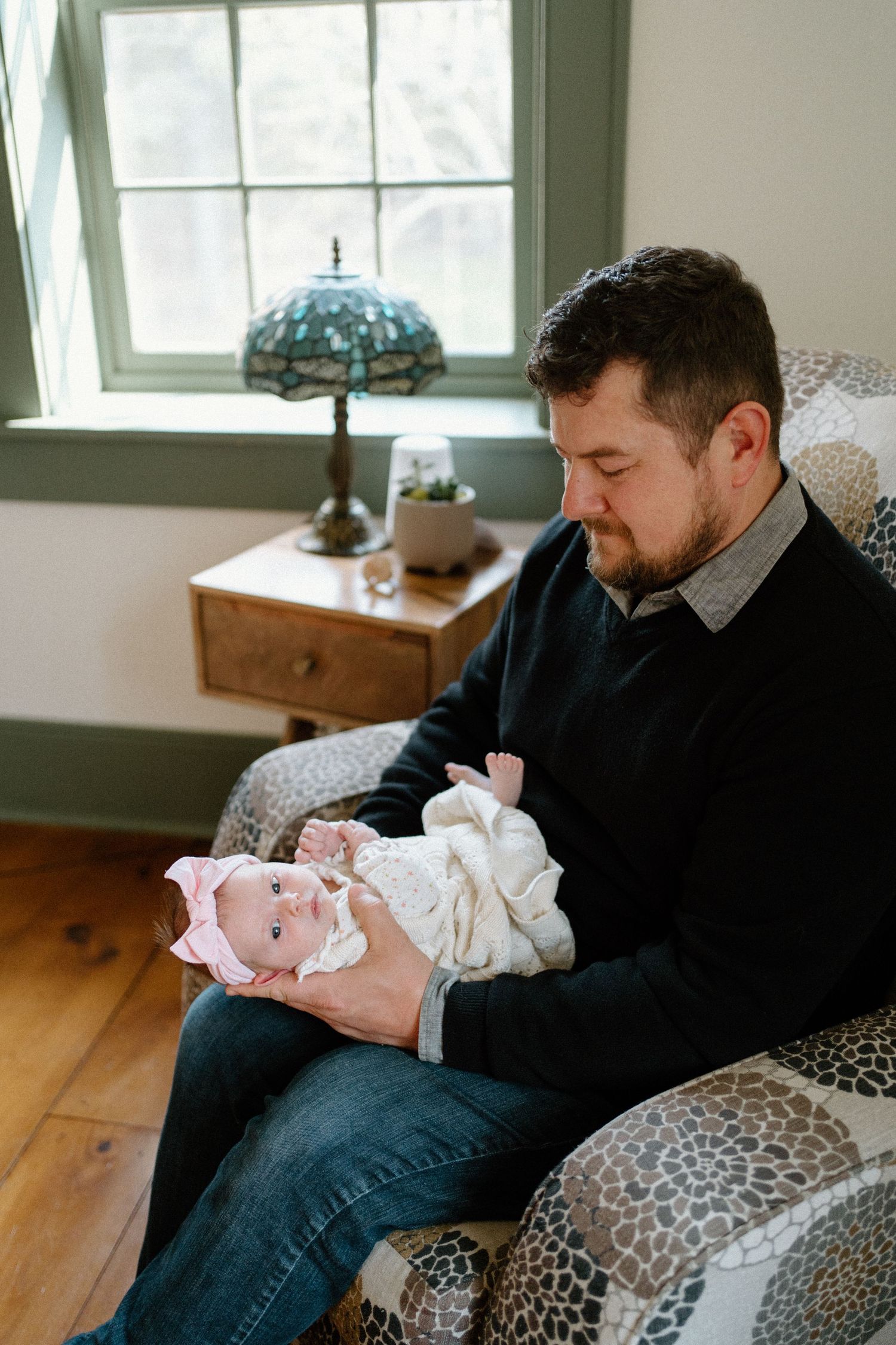 A tender moment as someone gently holds a swaddled newborn baby in a room with a vintage tiffany lamp and hardwood floors.