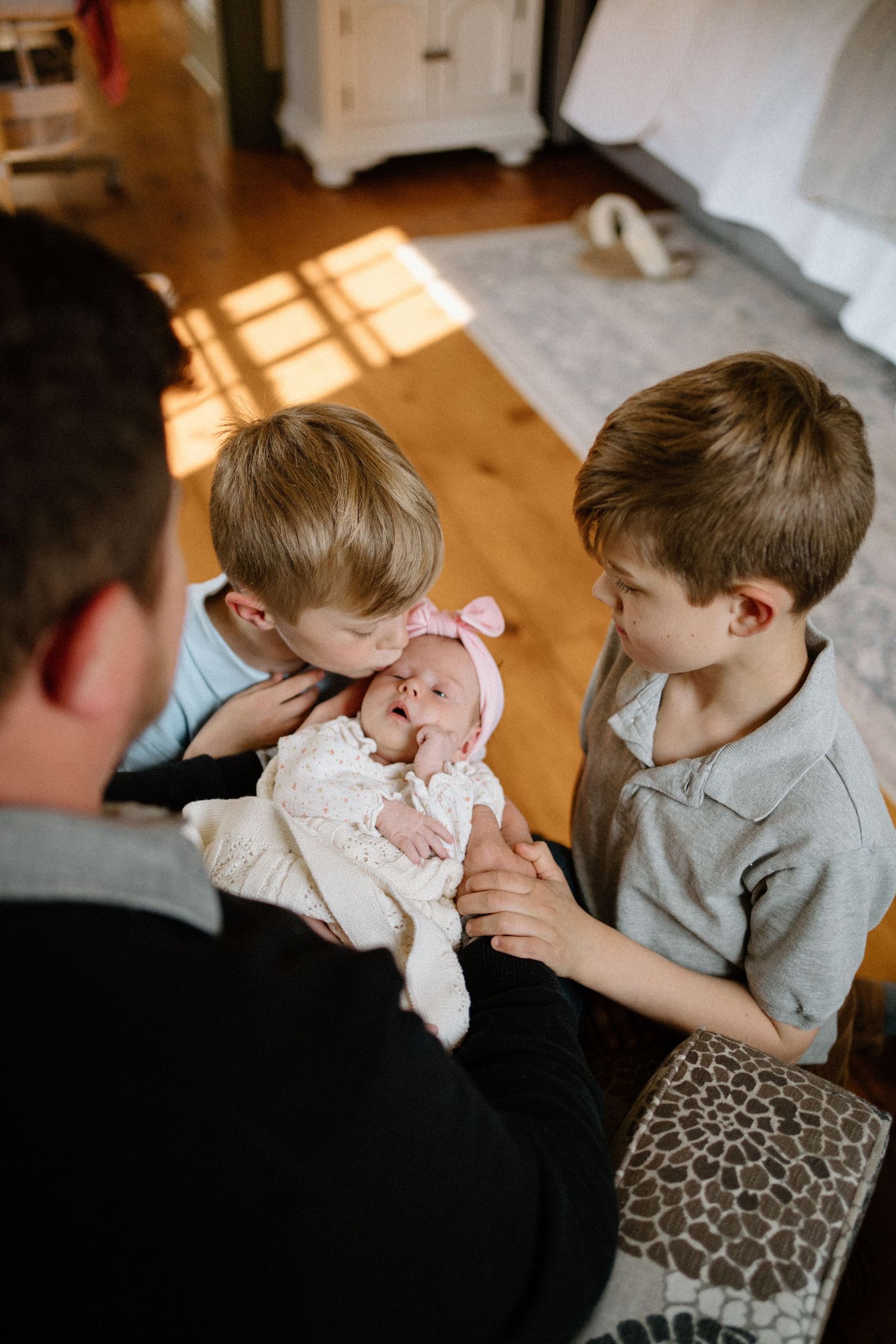 Two young boys lean in close to look at a newborn baby wearing a pink bow while sitting on a couch.