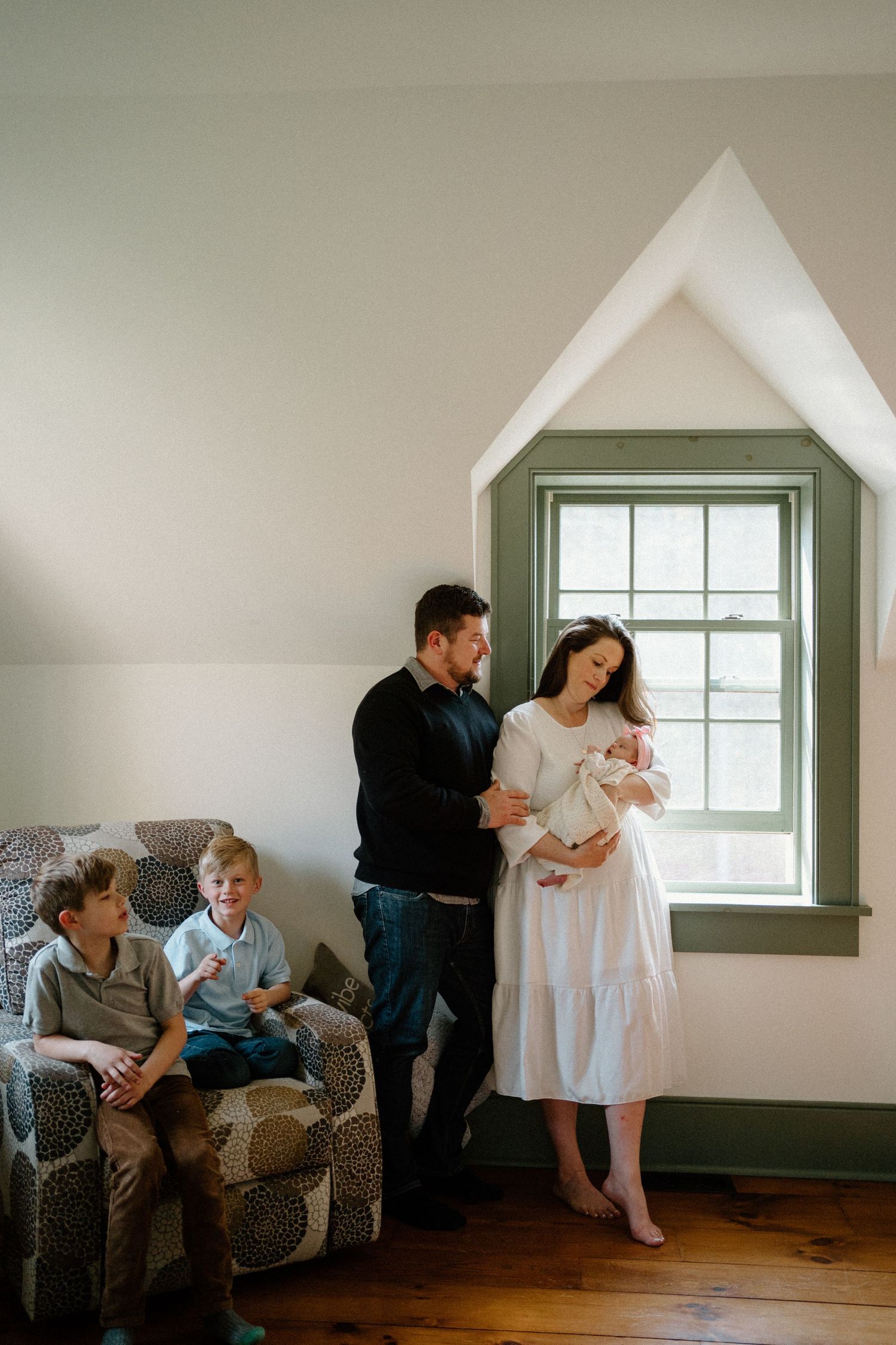 A family shares an intimate moment with a newborn baby in a bright room with a peaked window and hardwood floors.