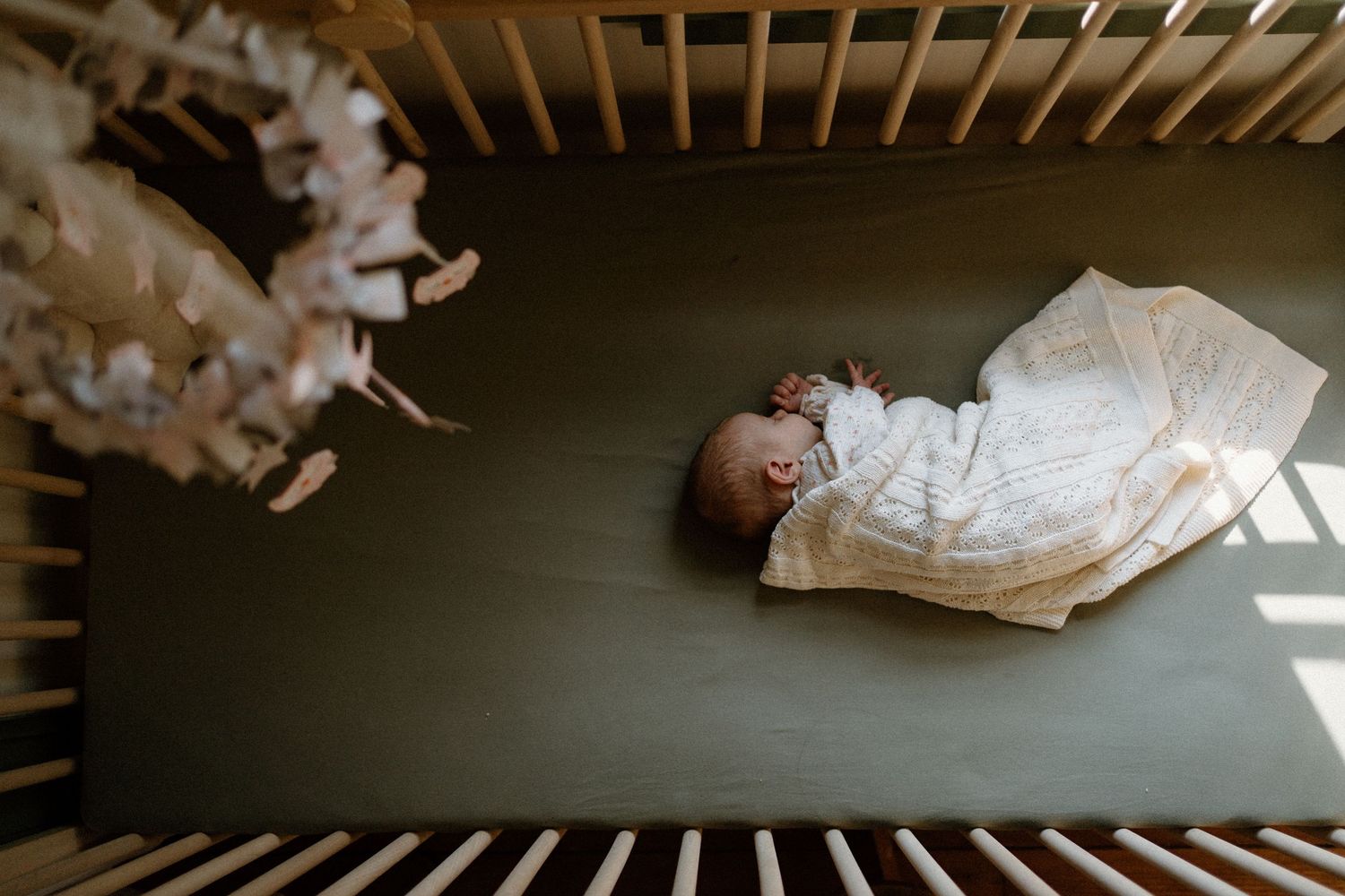 A baby wearing white clothing lies peacefully in a dark wooden crib viewed from above.