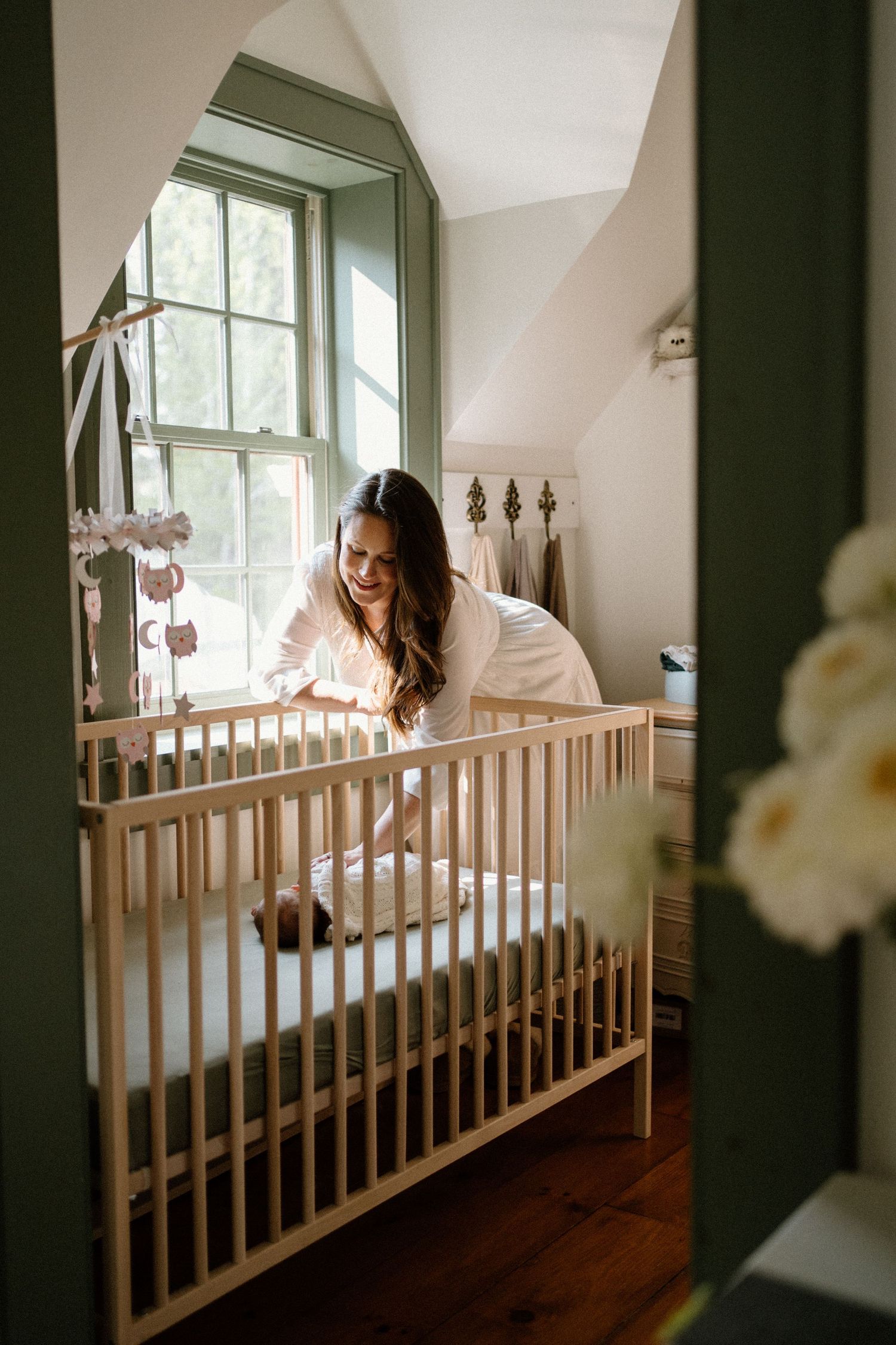A nursery scene with wooden crib by a bright window decorated with soft white flowers.
