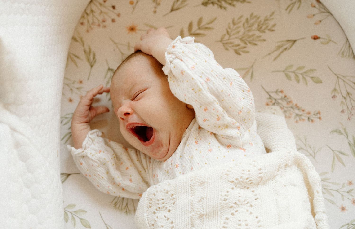 A yawning baby dressed in white lace clothing lies on a floral patterned surface.