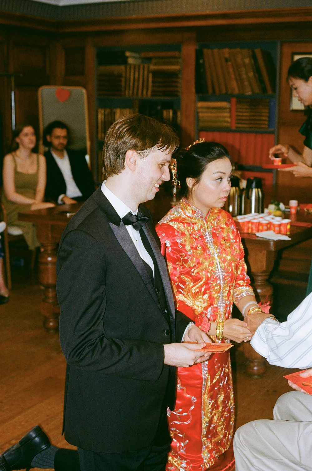 A couple at a wedding reception with the bride wearing a traditional red Chinese qipao dress and groom in black tuxedo.