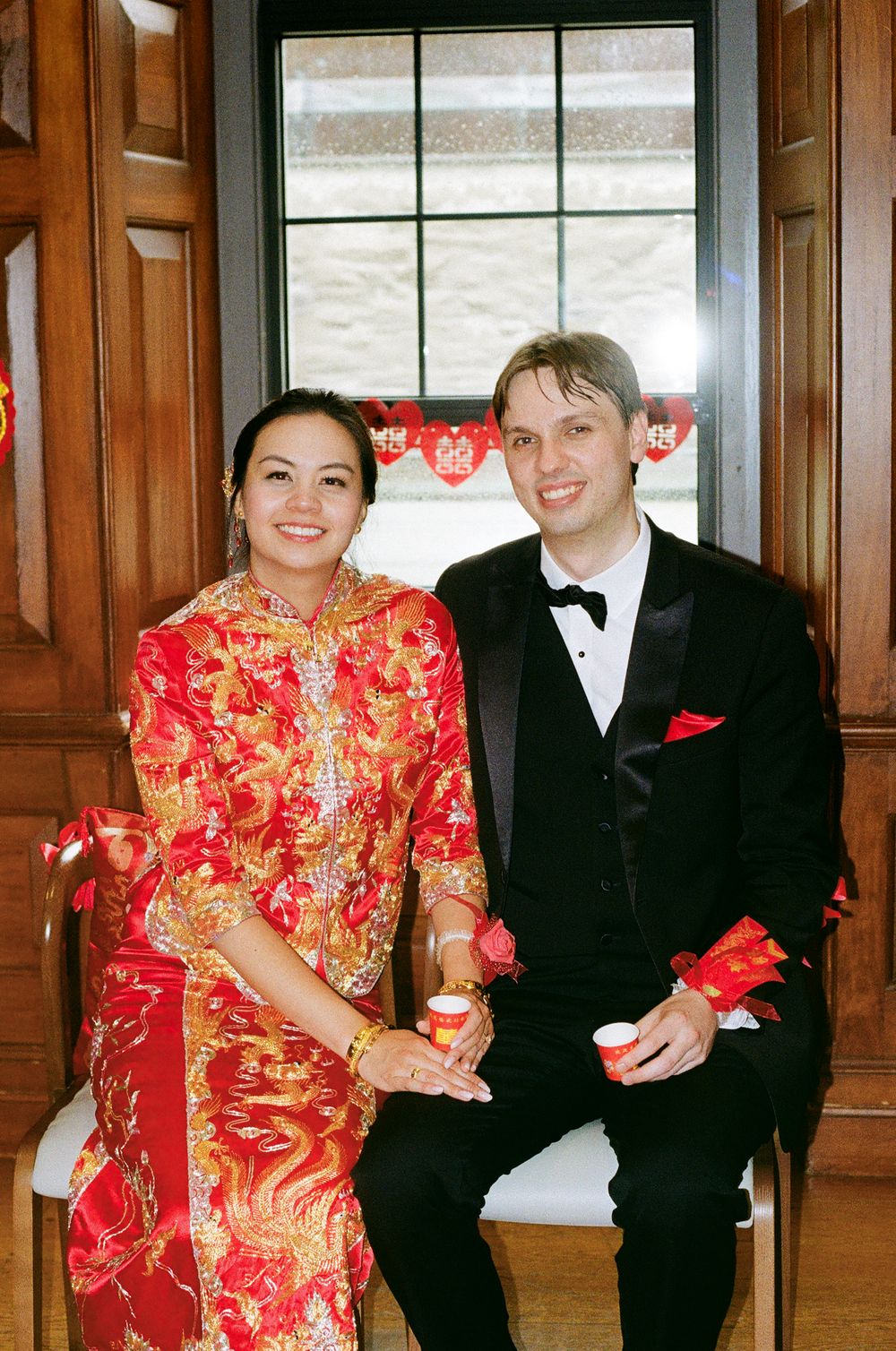 A bride in red Chinese wedding dress and groom in tuxedo pose by a window with red decorations.