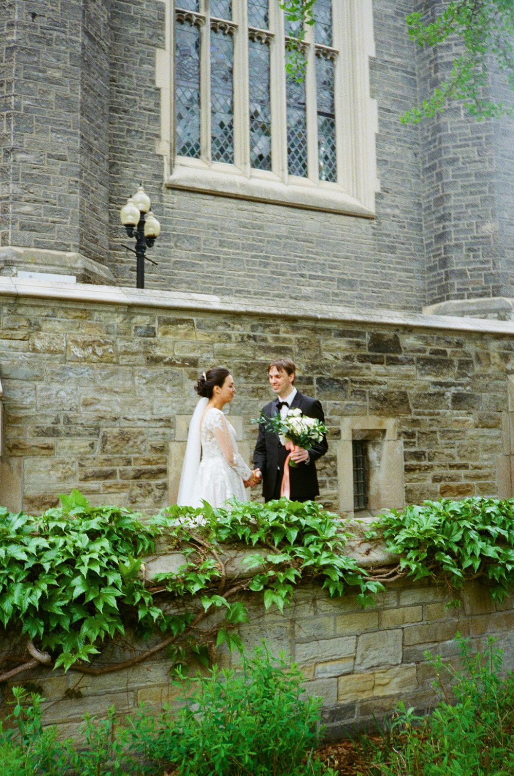 Stone church exterior with gothic windows and verdant landscaping creates romantic setting.