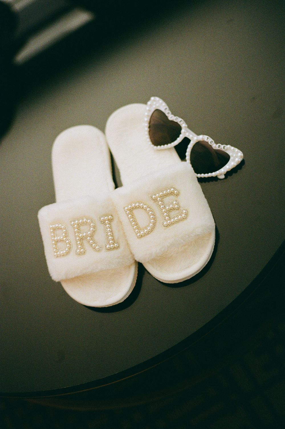White bride slippers with pearl lettering and heart-shaped sunglasses on dark surface.