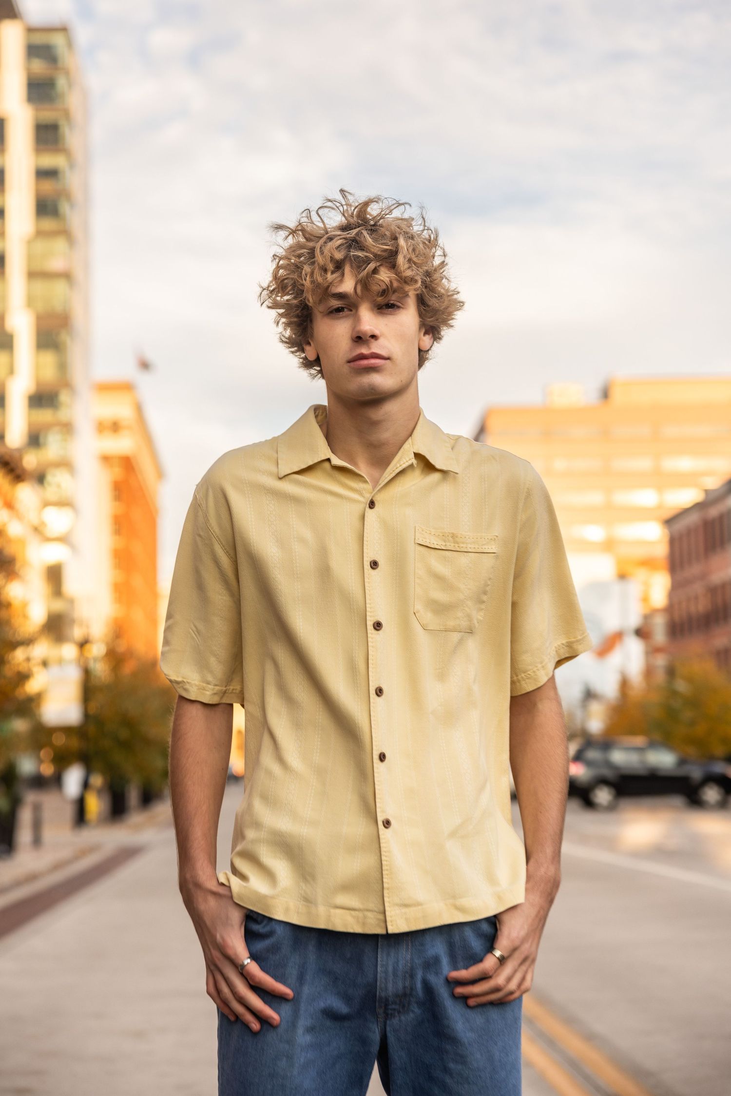 A person in a yellow short-sleeve button-up shirt and jeans stands on an urban street at sunset.