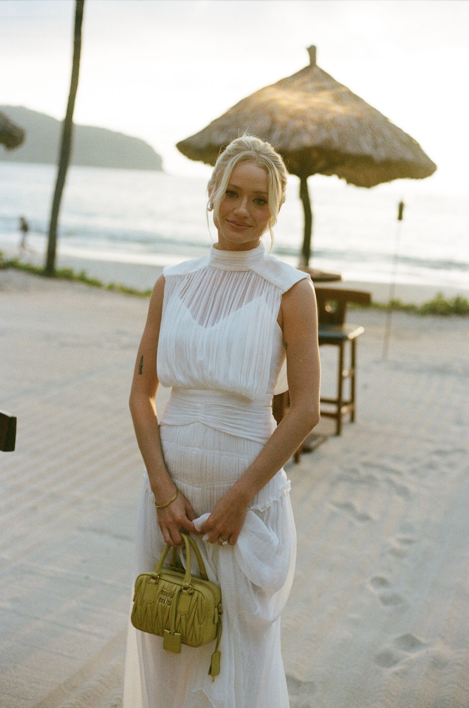 A woman in white dress poses on beach with thatched umbrellas and yellow handbag.
