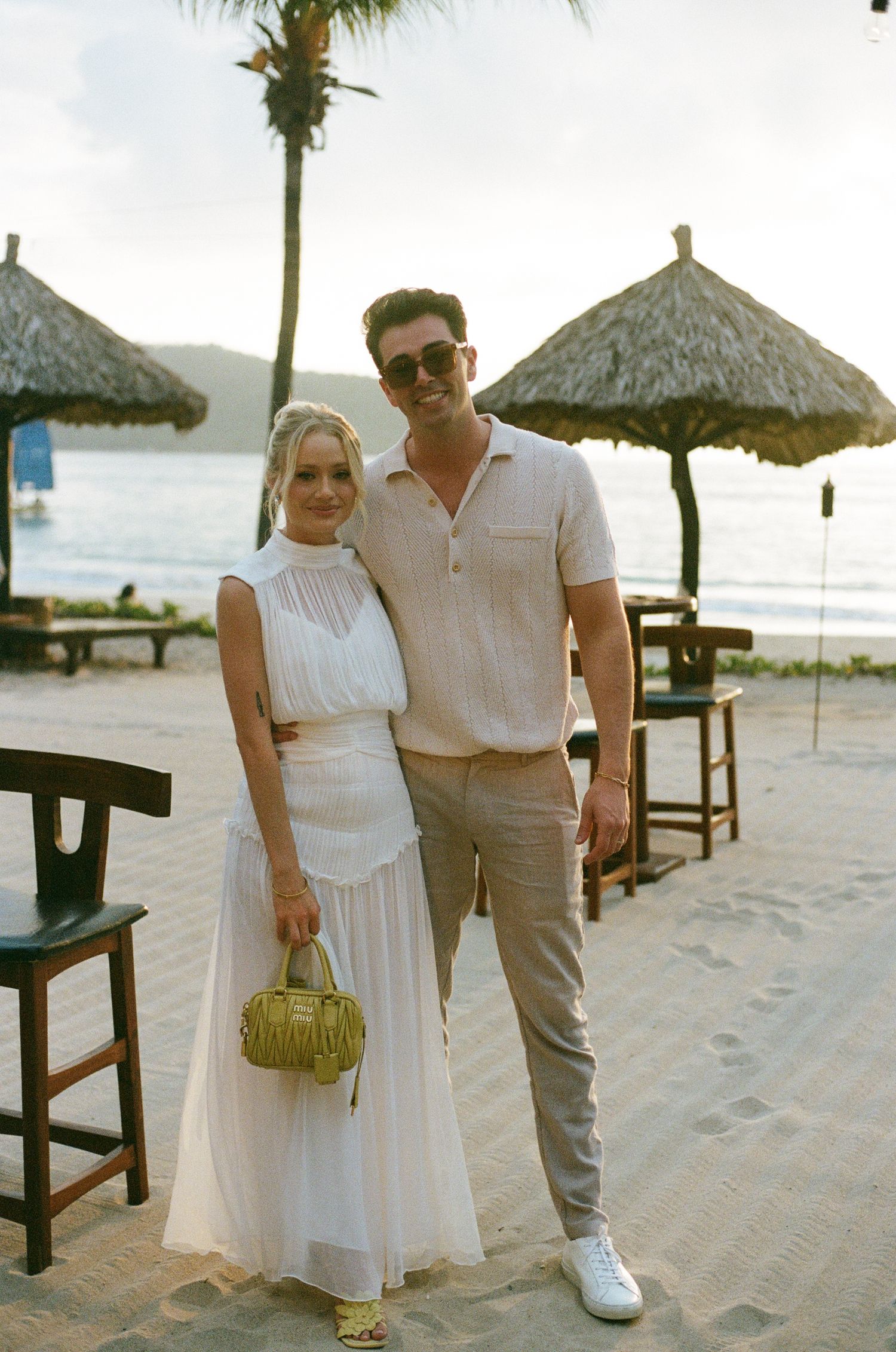 A stylish couple poses on beach with thatched umbrellas in background.