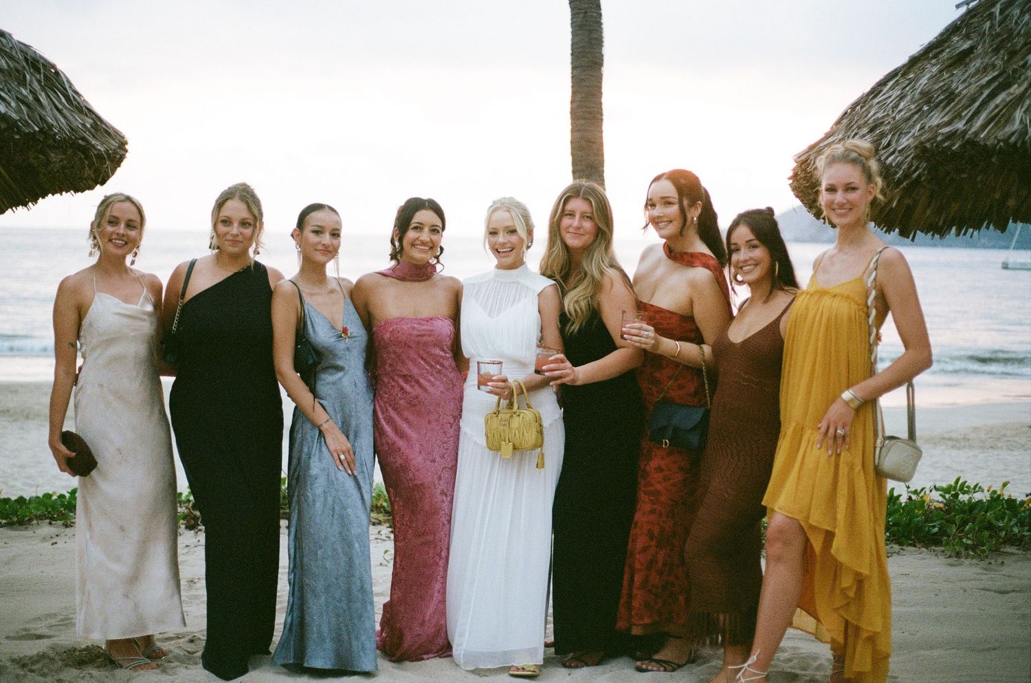 A group of bridesmaids in colorful dresses pose together on a beach at sunset.