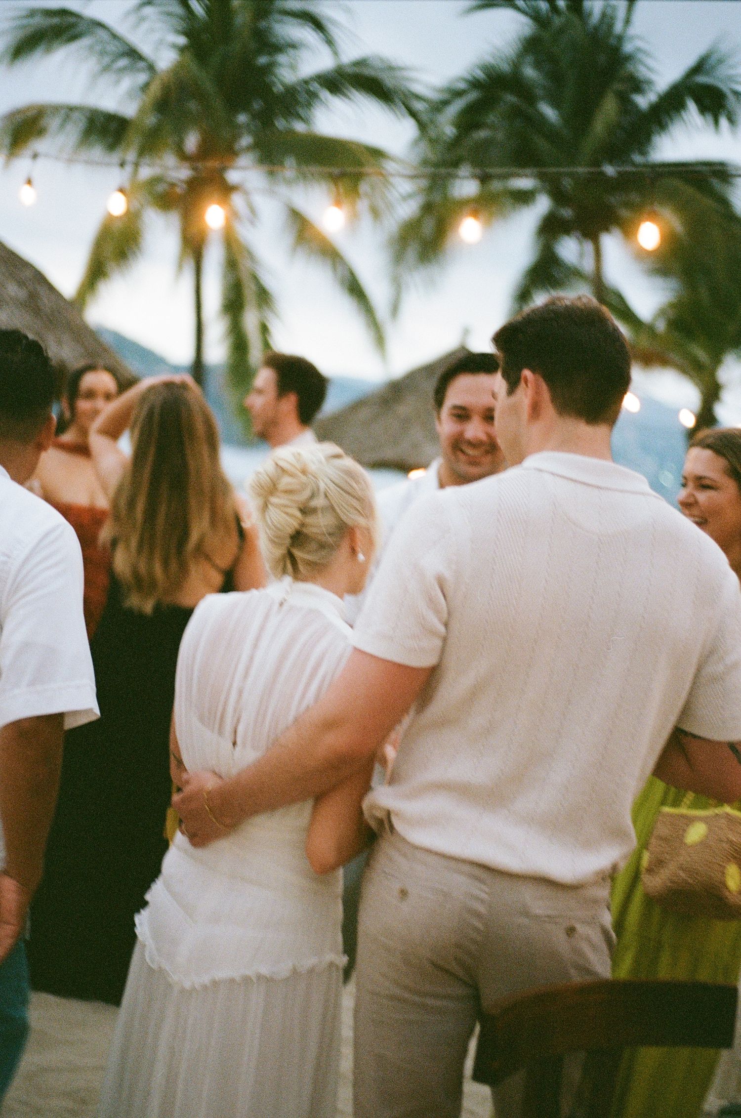 A beach wedding reception at sunset with palm trees and string lights creating a romantic tropical atmosphere.