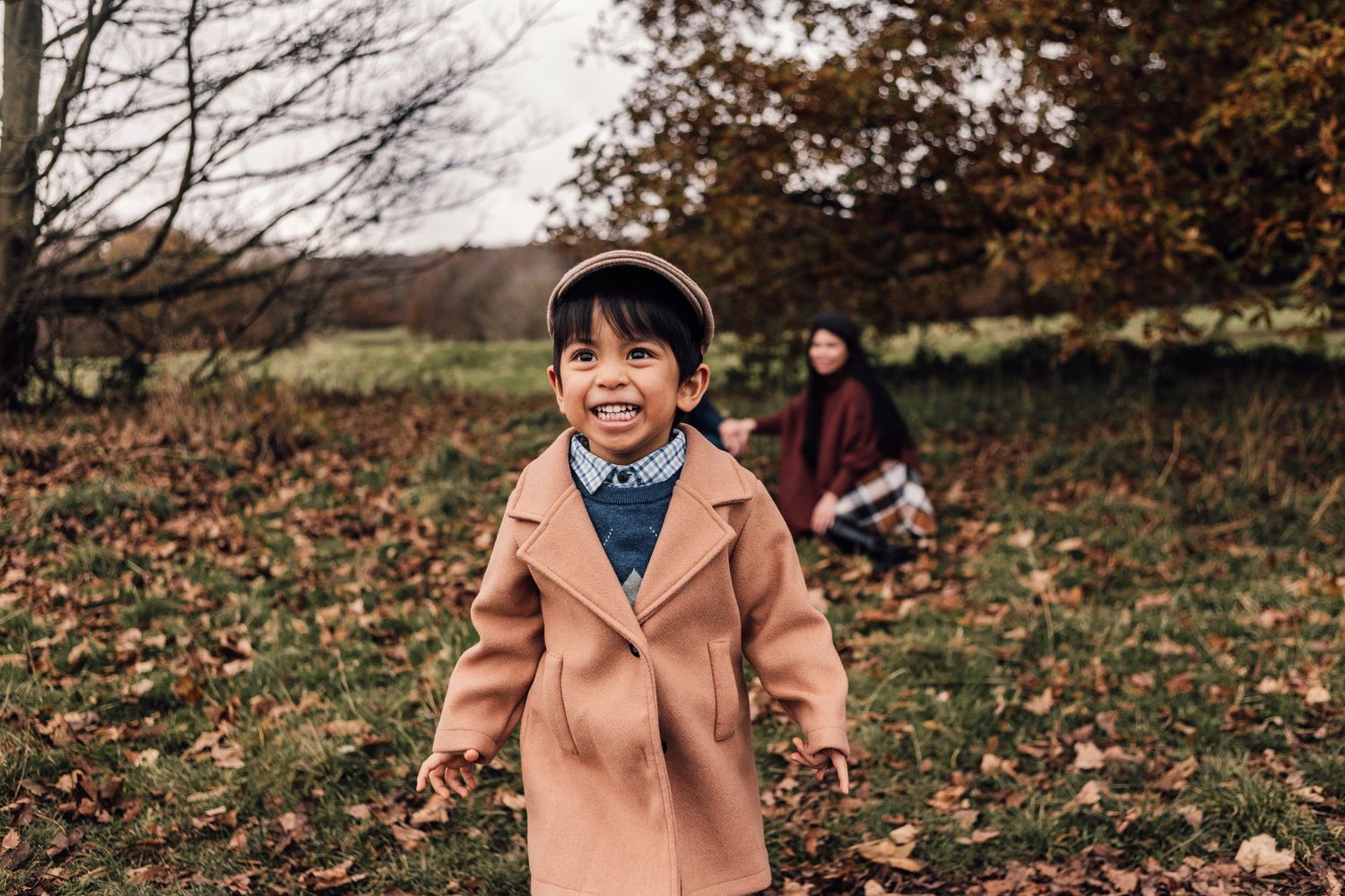 A child wearing a light brown peacoat smiles while playing in fallen autumn leaves with someone in the background.