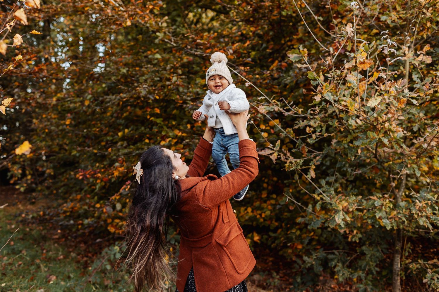 A person in a rust-colored coat lifts a small child wearing a winter hat among autumn foliage.
