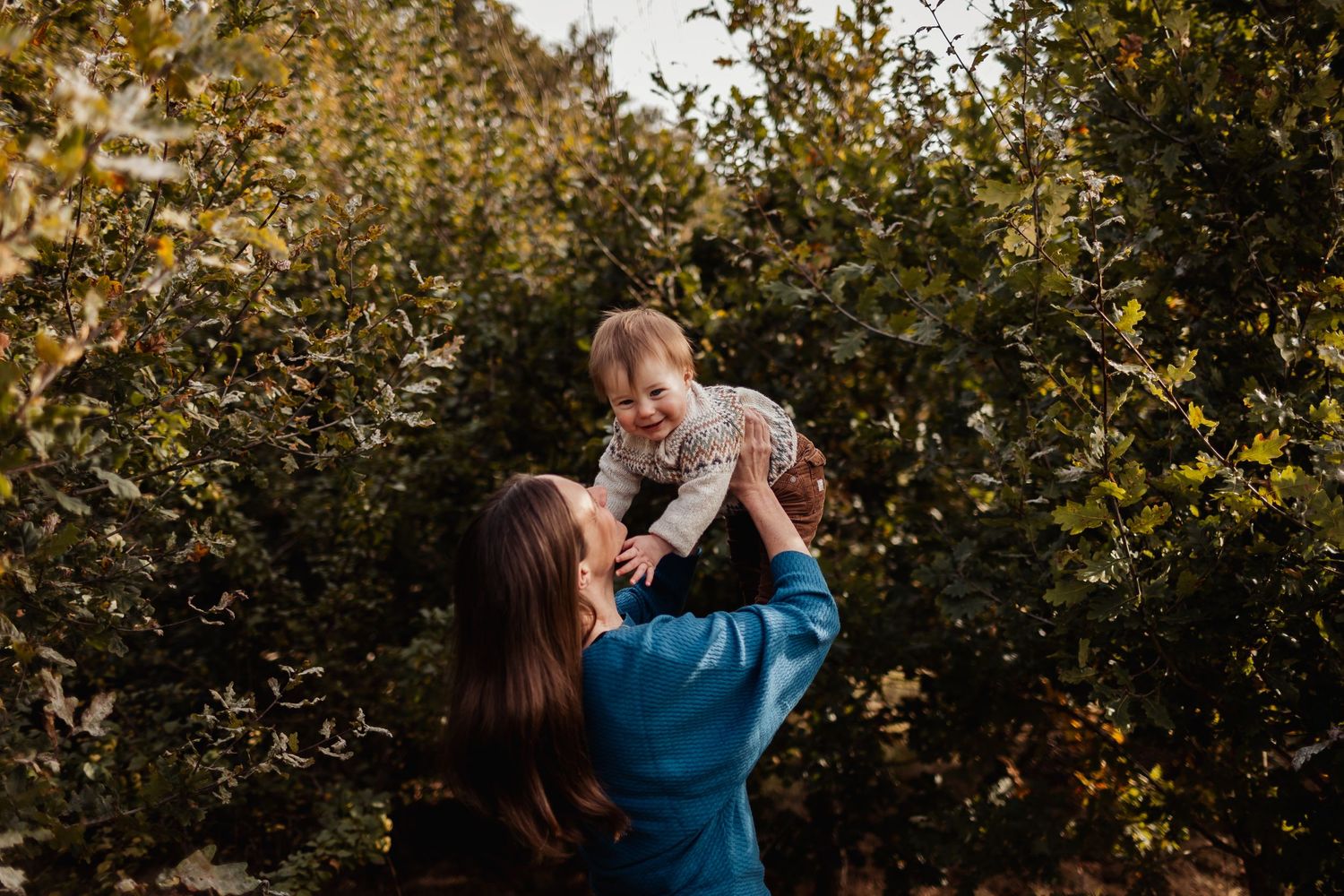 Parent lifts child up joyfully in a sunlit orchard during golden hour.