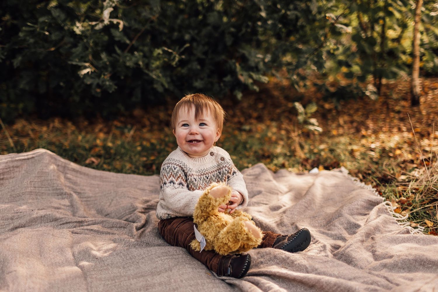A smiling toddler sits on a blanket outdoors with a teddy bear toy.