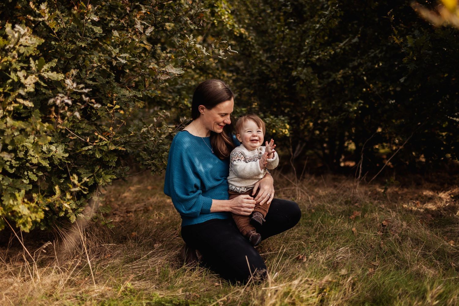 An adult in a blue sweater holds a small child outdoors in an autumn wooded setting.