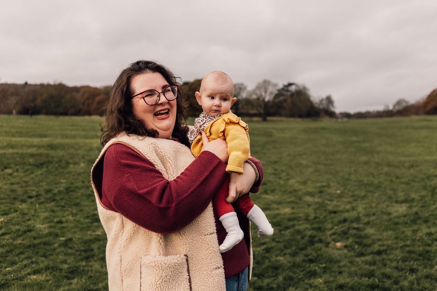 A person in a burgundy and beige sweater holds a baby wearing yellow on a grassy field during an overcast day.