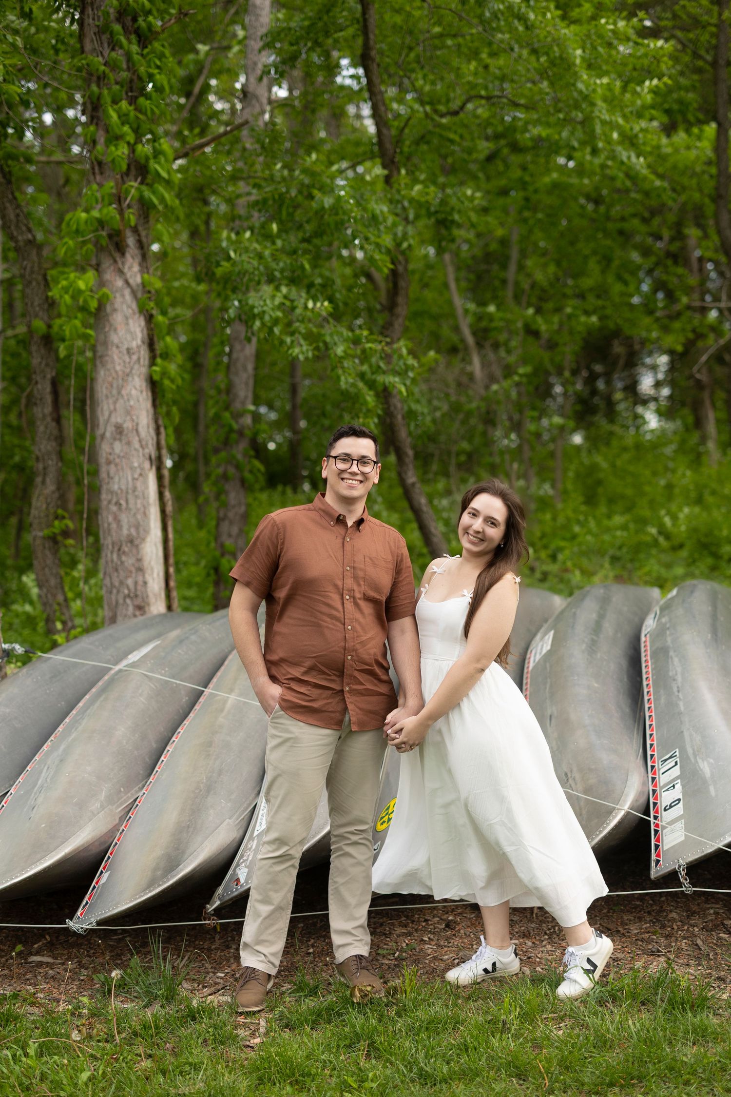 A couple poses outdoors in casual attire next to upturned canoes on a grassy lawn surrounded by green trees.