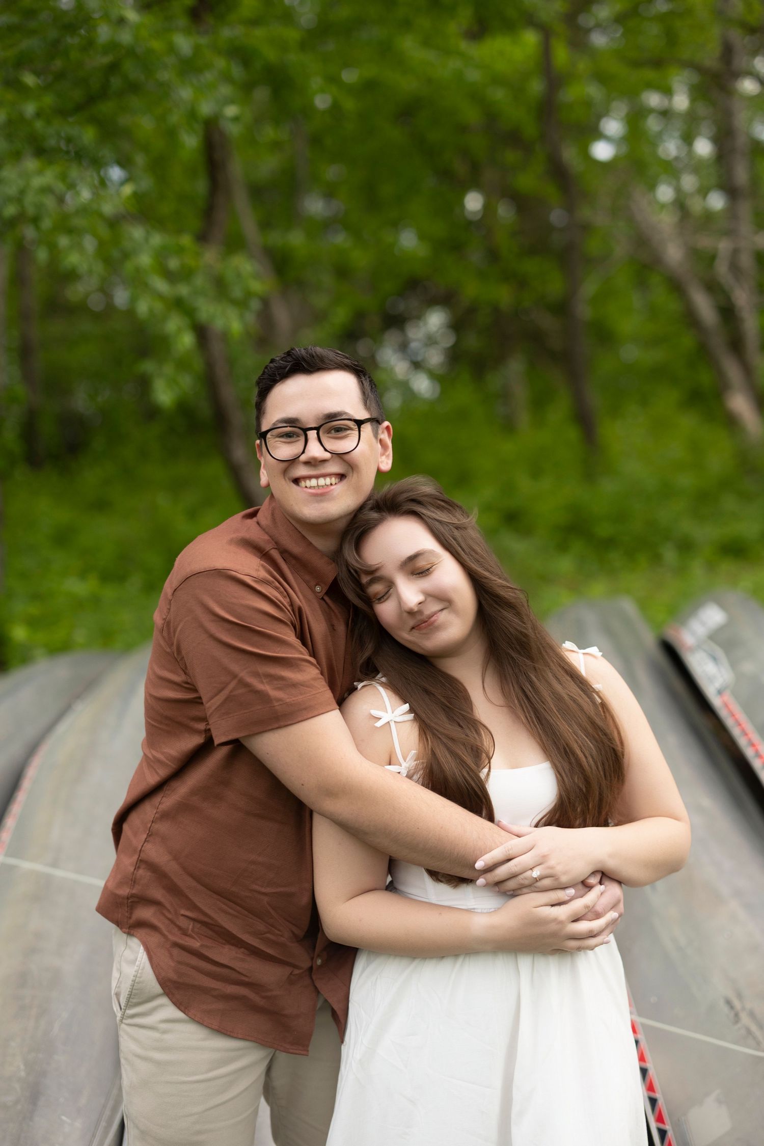 A couple embraces while standing on wooden planks outdoors with lush green trees in the background.