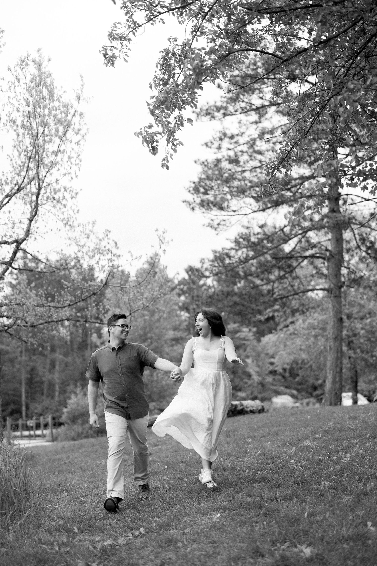 Couple enjoying a romantic black and white moment while walking together on a grassy hill with trees in the background.