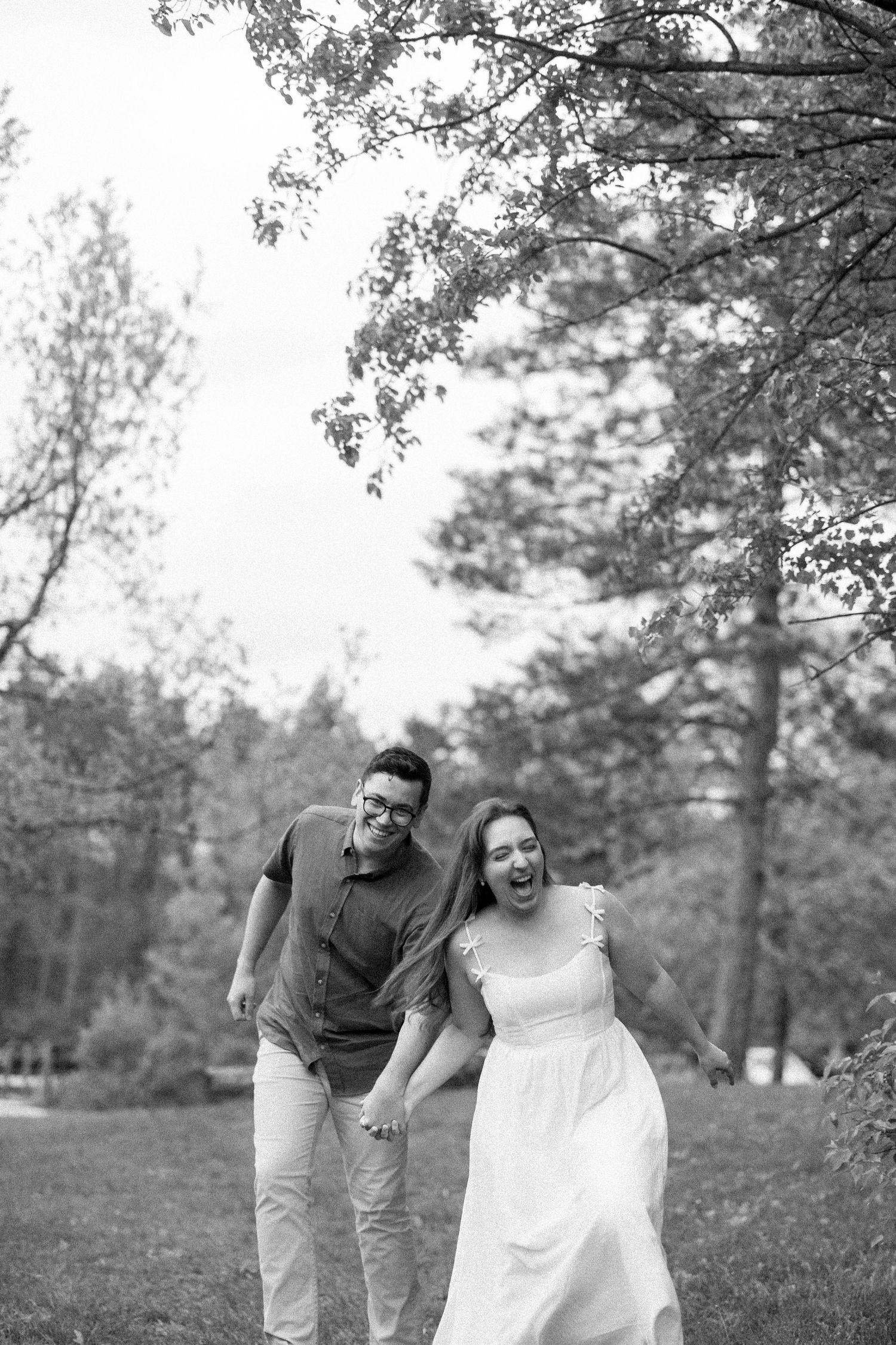 A joyful couple laughs together while walking hand in hand through a scenic wooded park in black and white.