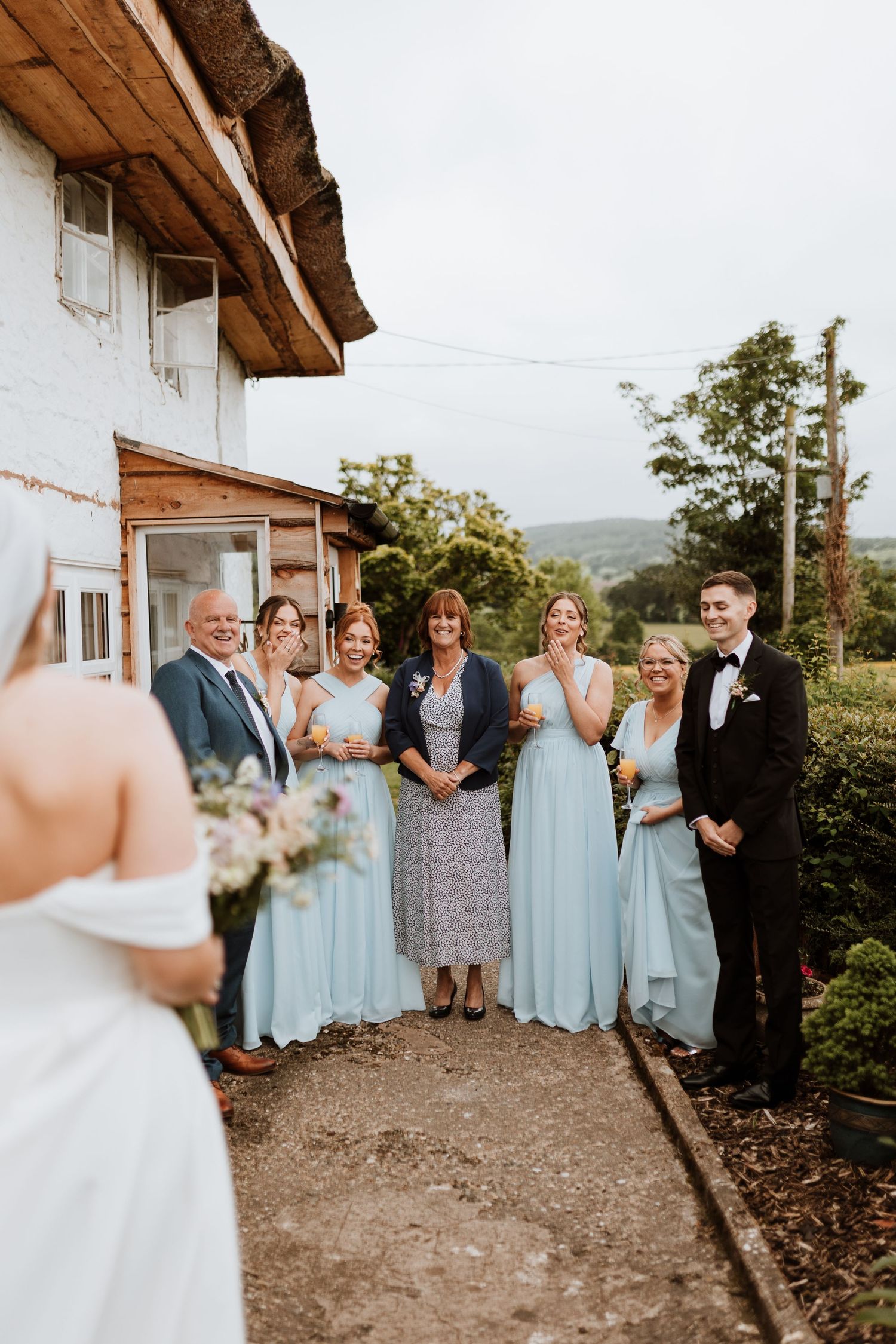 Wedding party in light blue bridesmaid dresses gathered outside a rustic white building with mountain views.