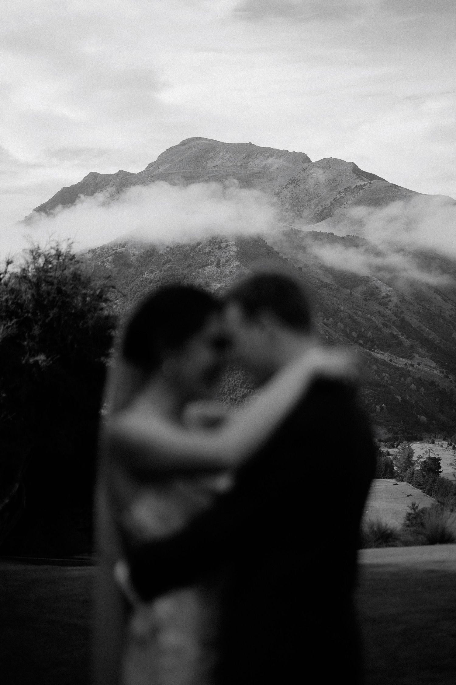 A romantic moment silhouetted against misty mountain peaks in a moody black and white landscape.