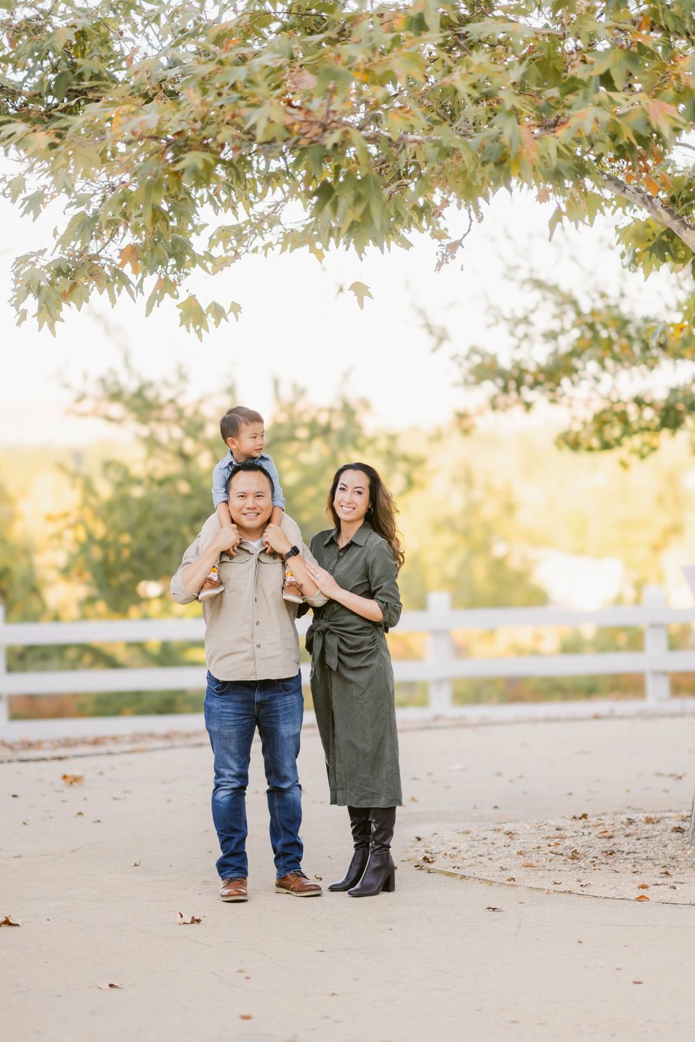 A family enjoys a warm autumn day outdoors under a tree canopy with fall foliage and a white fence in the background.
