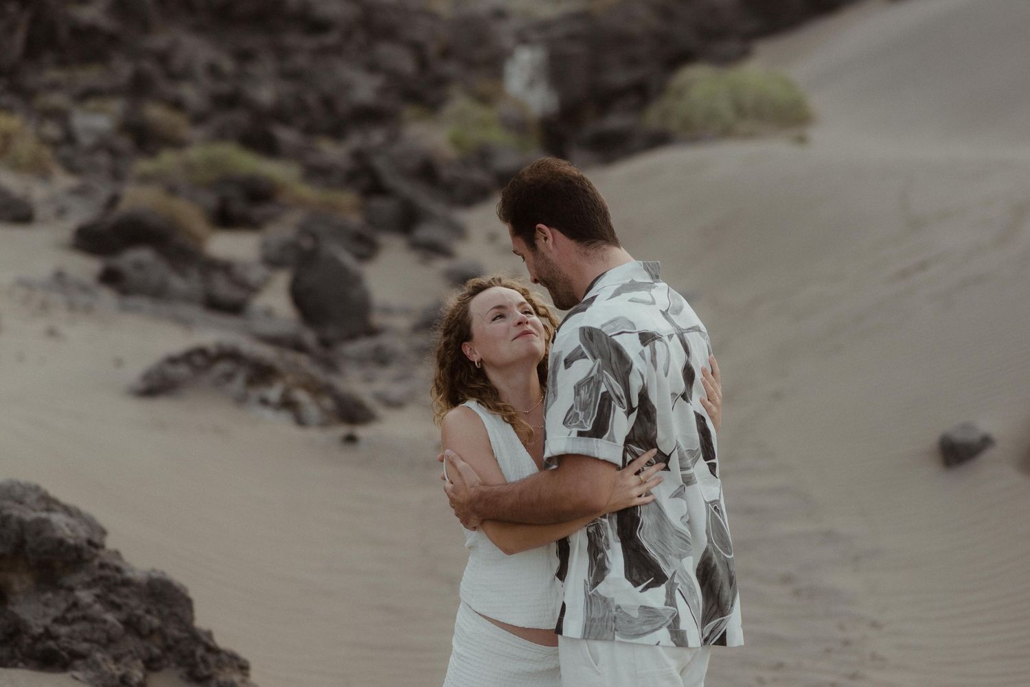 Couple embraces on a sandy beach with volcanic rocks in the background during a romantic outdoor photoshoot.