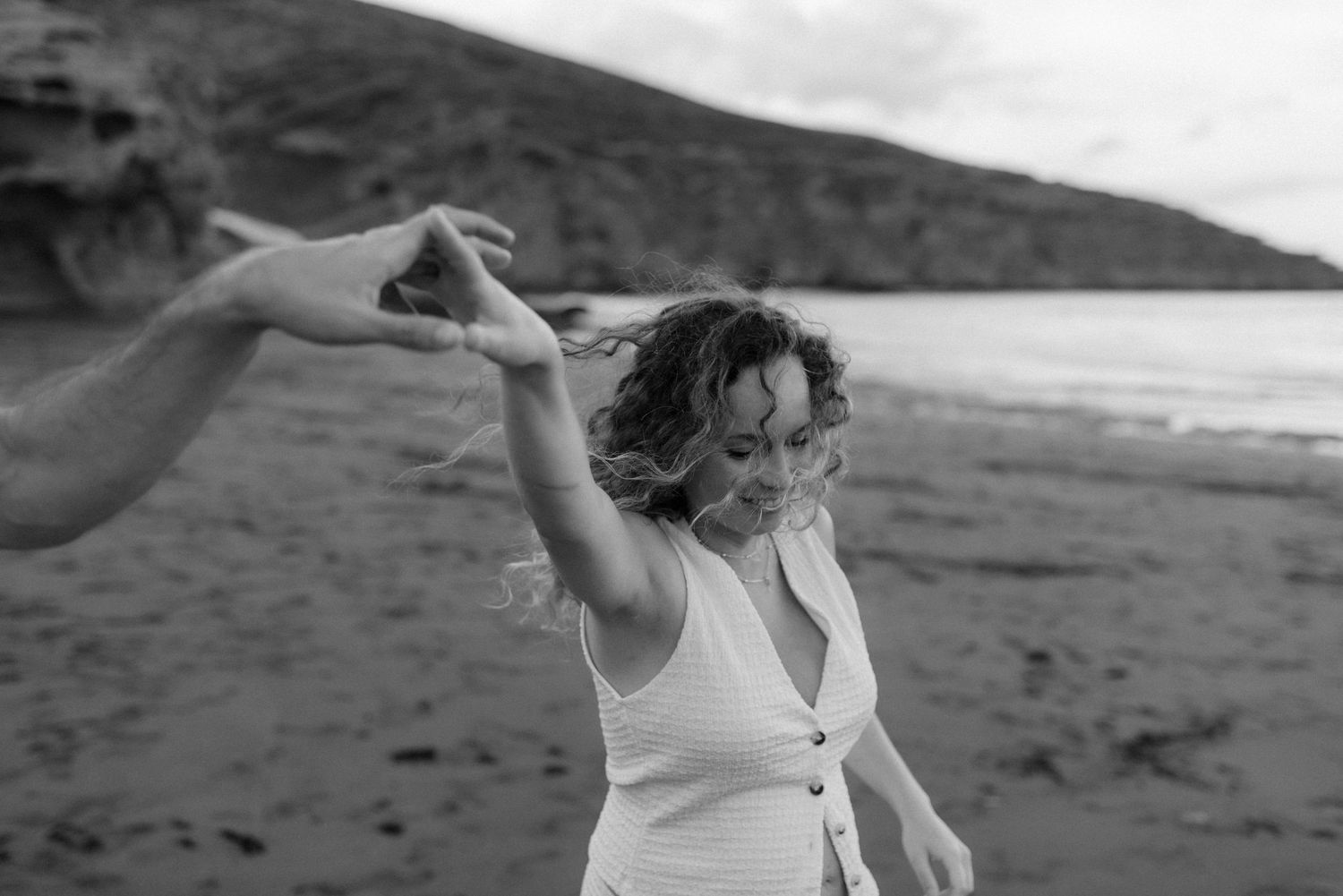Black and white photo shows a person in a white dress dancing joyfully on a beach with hills in the background.