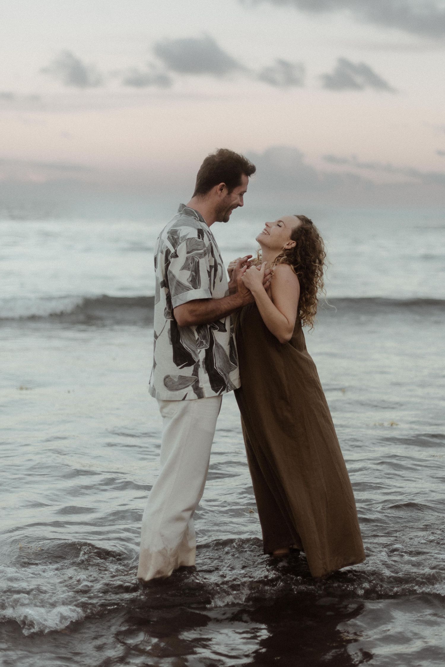 A romantic couple embraces in the ocean waves at sunset wearing casual beachwear.