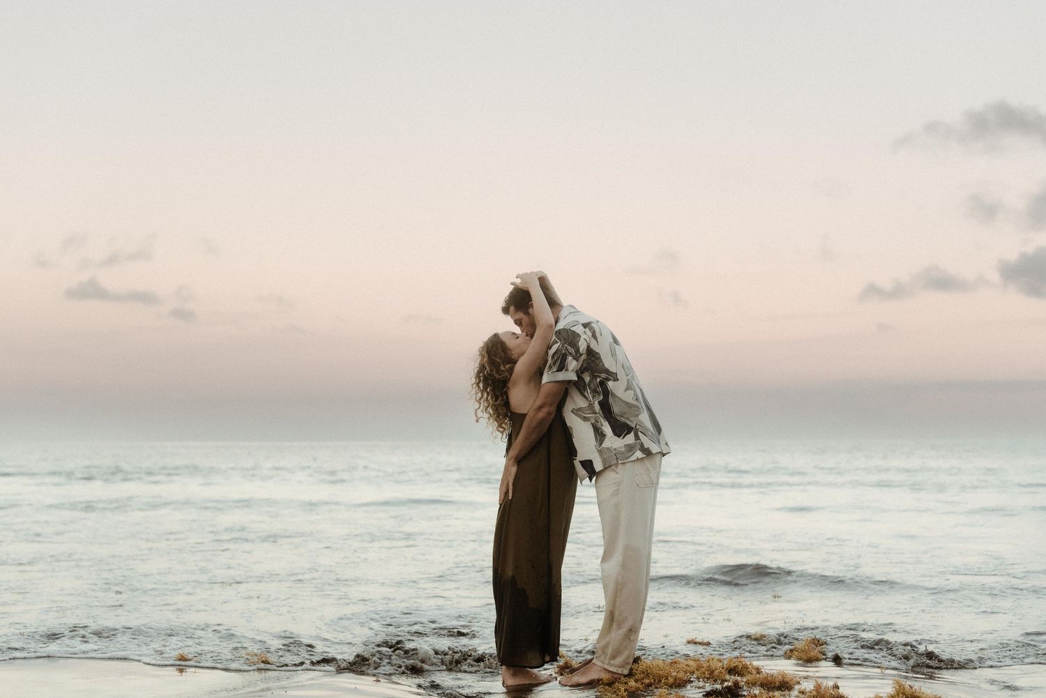 Two people embrace tenderly on rocky beach during sunset with ocean waves and pink sky in background.