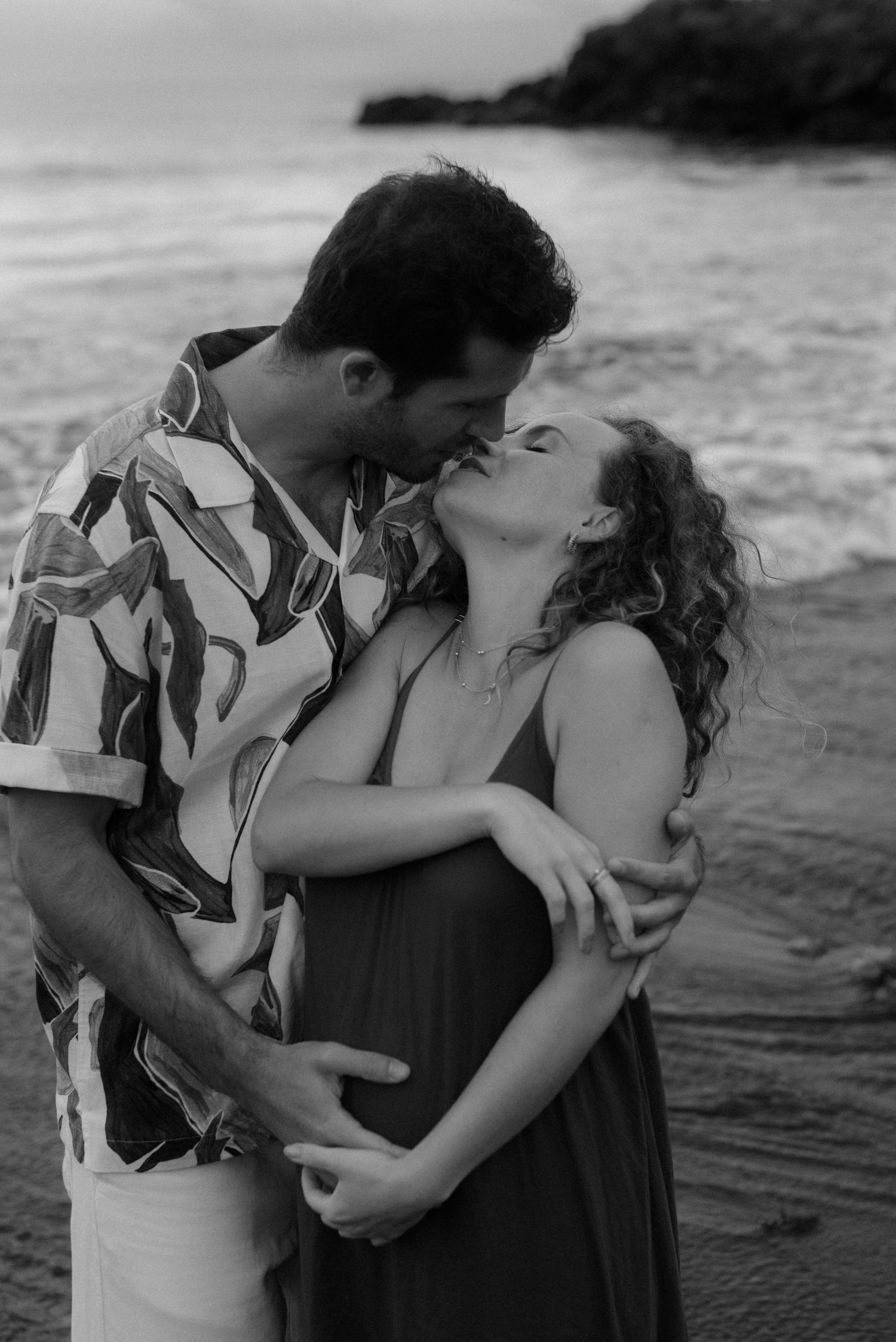 A romantic couple embraces at the beach in black and white as waves crash behind them at sunset along the shore.
