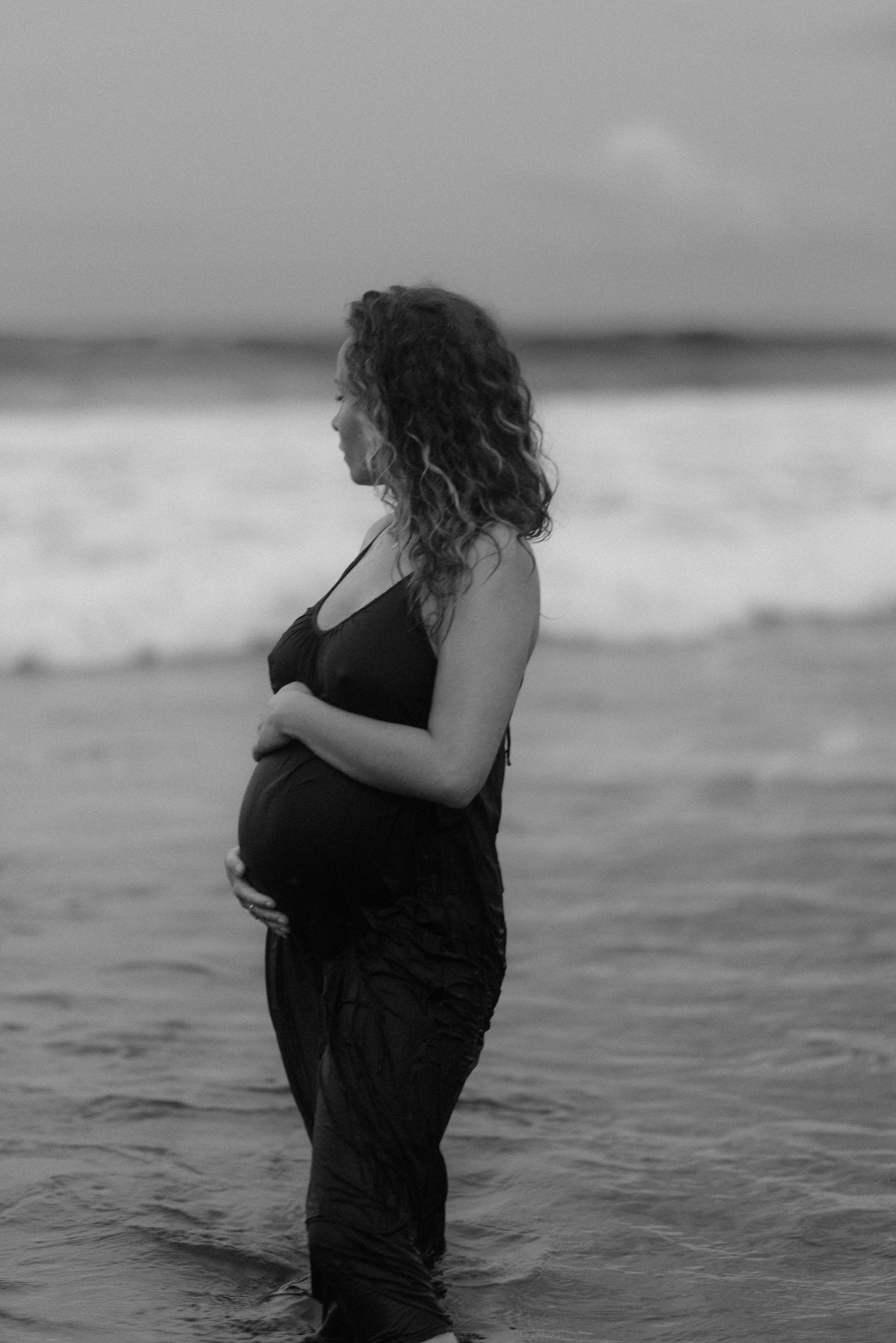 A pregnant woman stands in ocean waves wearing a black dress in a moody black and white photograph.
