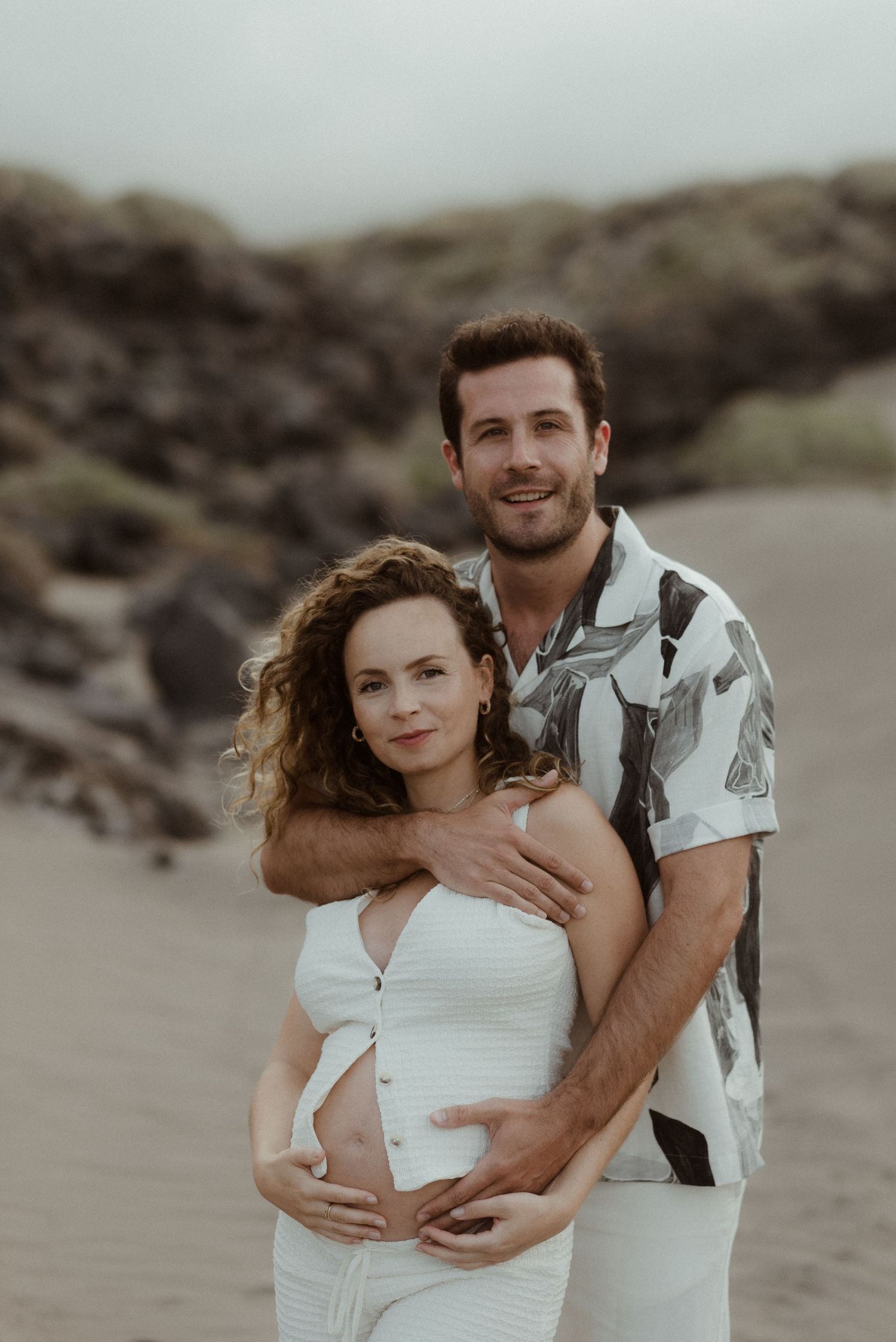 Romantic couple embracing on a beach in casual white outfits during an intimate photoshoot at sunset.