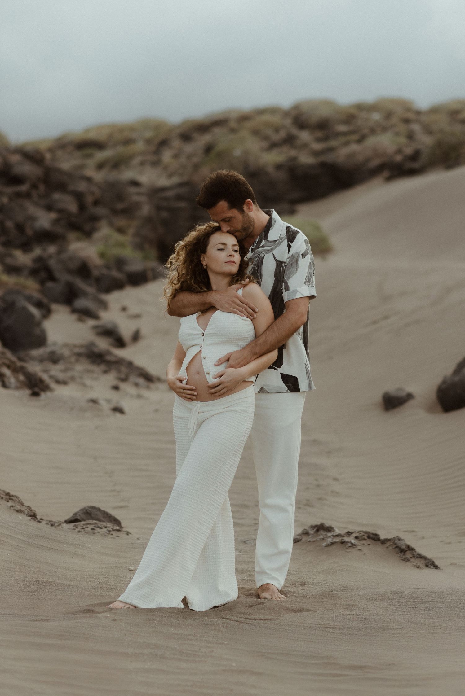 A romantic beach maternity photo featuring a couple embracing on sandy dunes at sunset with dramatic rocky outcrops.