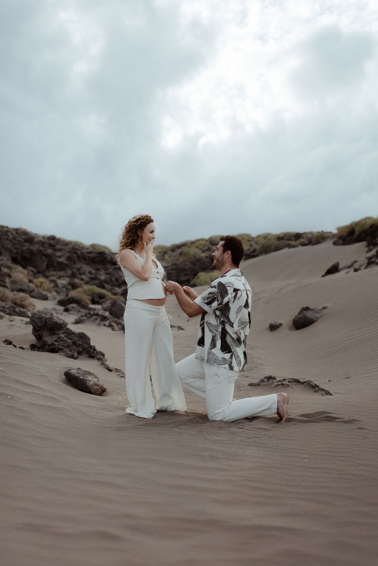 A romantic marriage proposal scene on a sandy beach with dramatic rocky cliffs and cloudy skies in the background.
