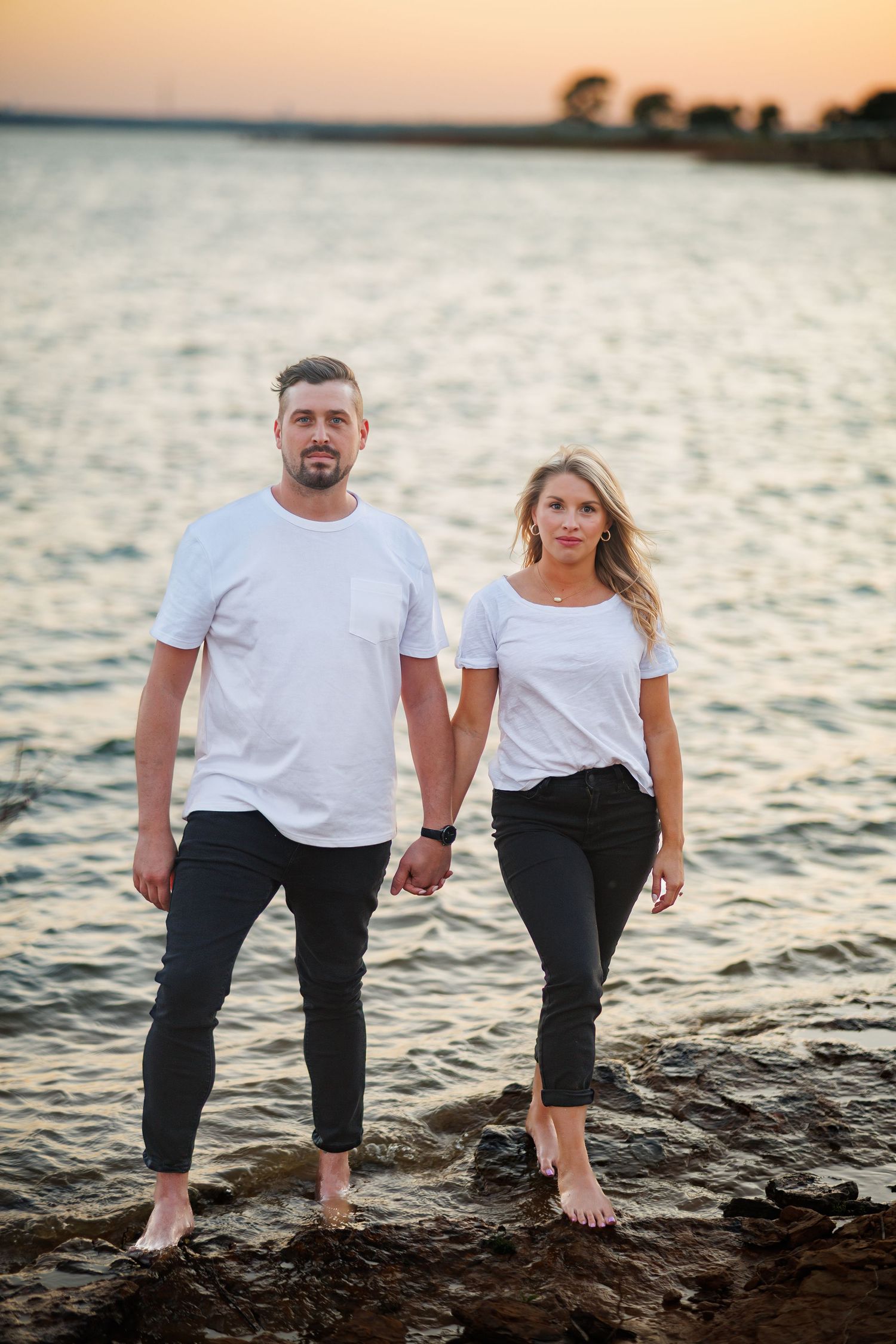 A couple in matching white t-shirts holding hands while standing barefoot at Rockwall Harbot in Dallas at sunset