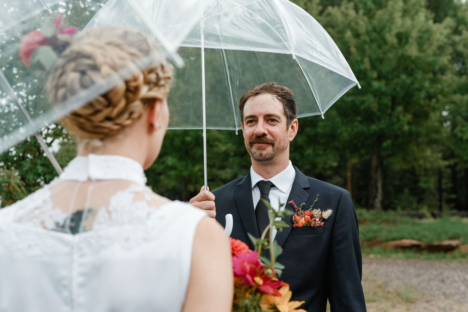 A couple shares a tender moment under a clear umbrella during an outdoor wedding in nature.