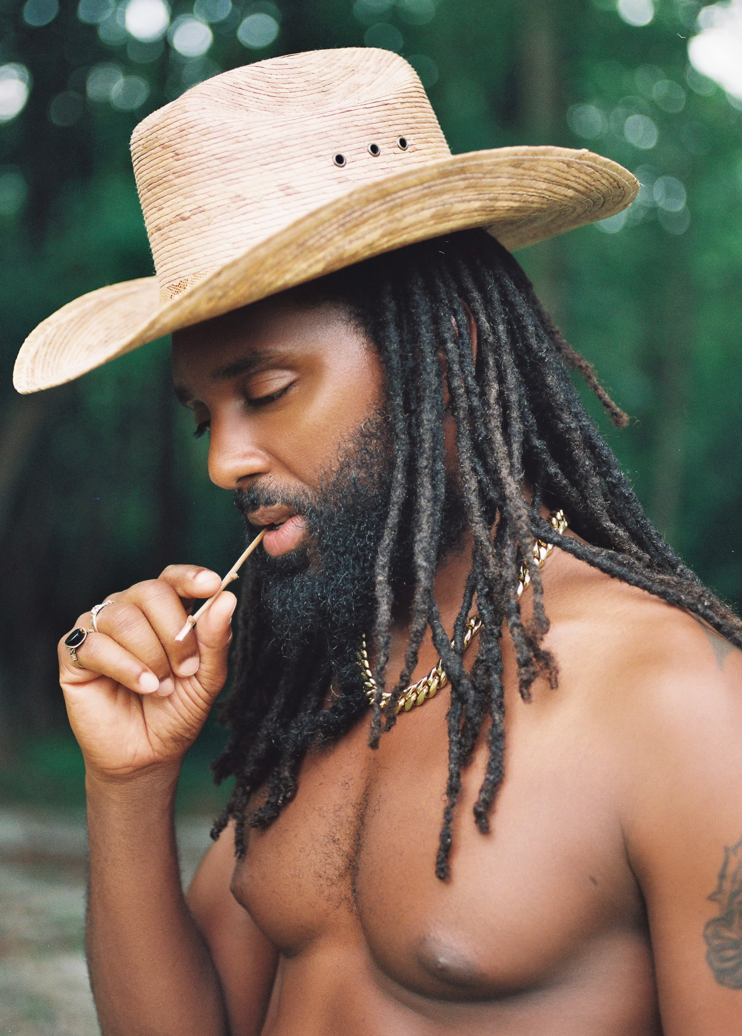 A person wearing a straw cowboy hat and dreadlocks poses shirtless against a blurred green outdoor backdrop.