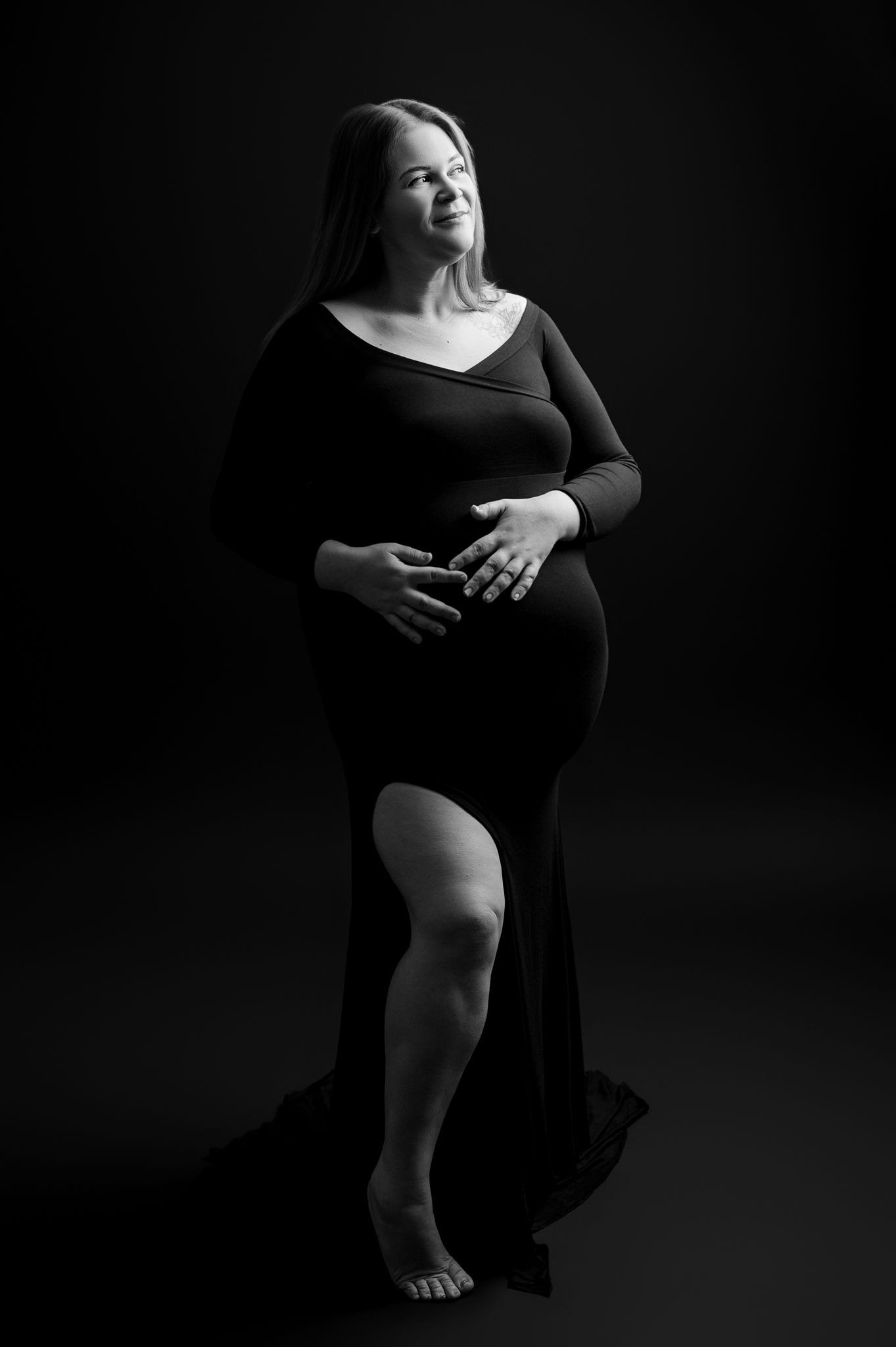 Black and white artistic maternity portrait of figure in flowing dress against dark background, highlighting graceful silhouette.