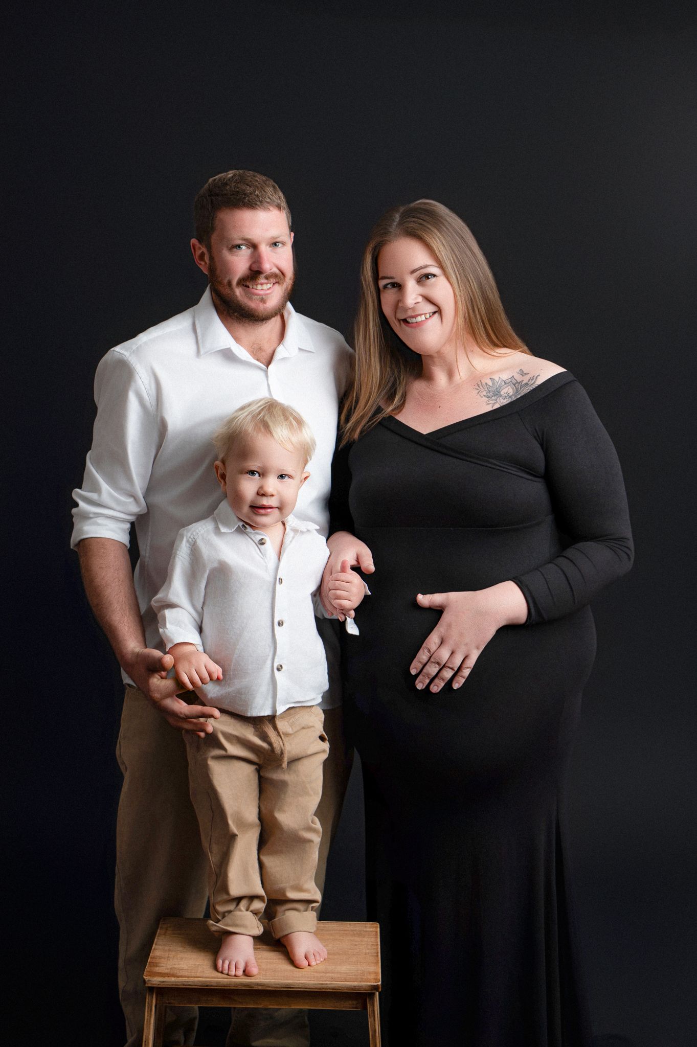 A couple with a toddler in matching white shirts pose against a black backdrop for a series of maternity photos.