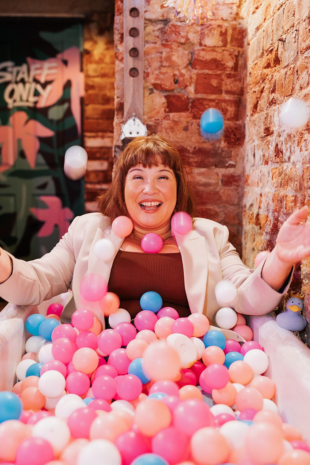 A joyful person tosses colourful pink, white and blue balls in the air while sitting in a bathtub against a brick wall.