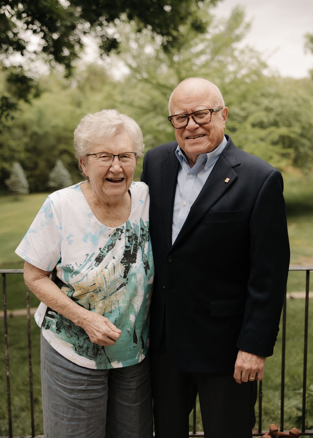 Two seniors sharing a happy moment together outdoors on a porch railing with trees in the background.