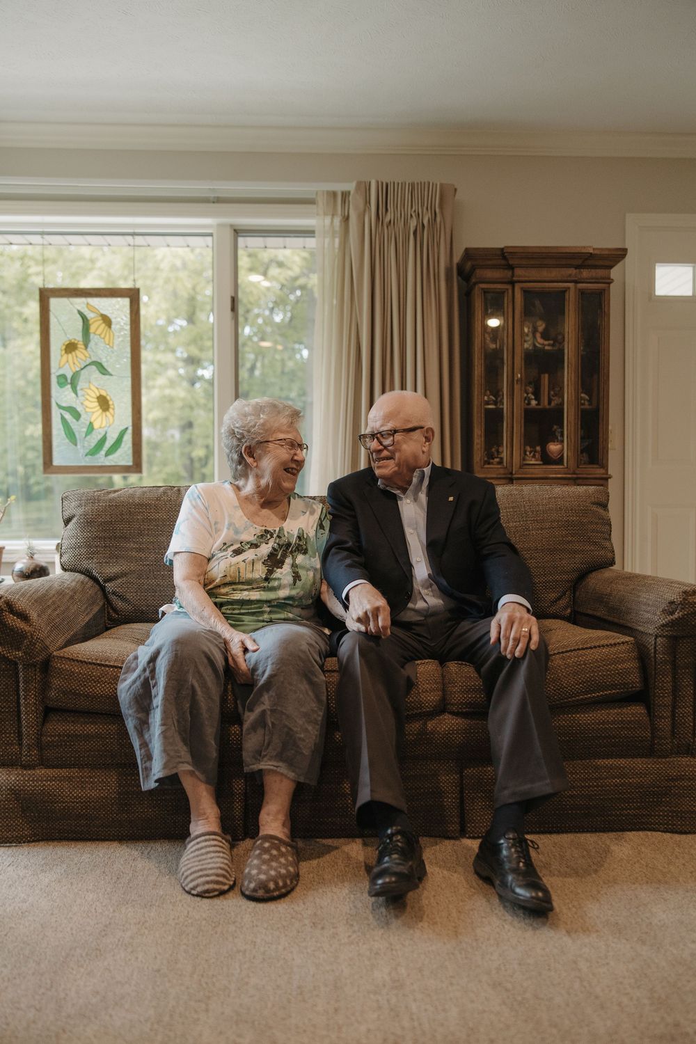 An elderly couple sits together on a brown leather couch in a bright living room with floral artwork on the walls.