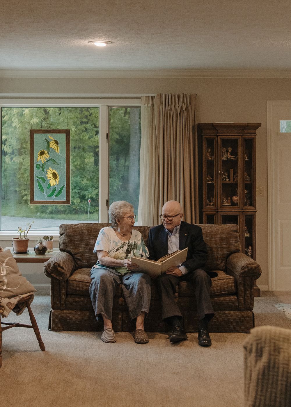 Two figures seated on a couch share a quiet moment together in a cozy living room with wood furniture and natural light.