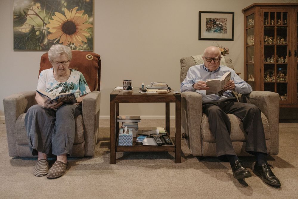 Two elderly people sit in armchairs reading books in a cozy living room with a wooden cabinet in the background.