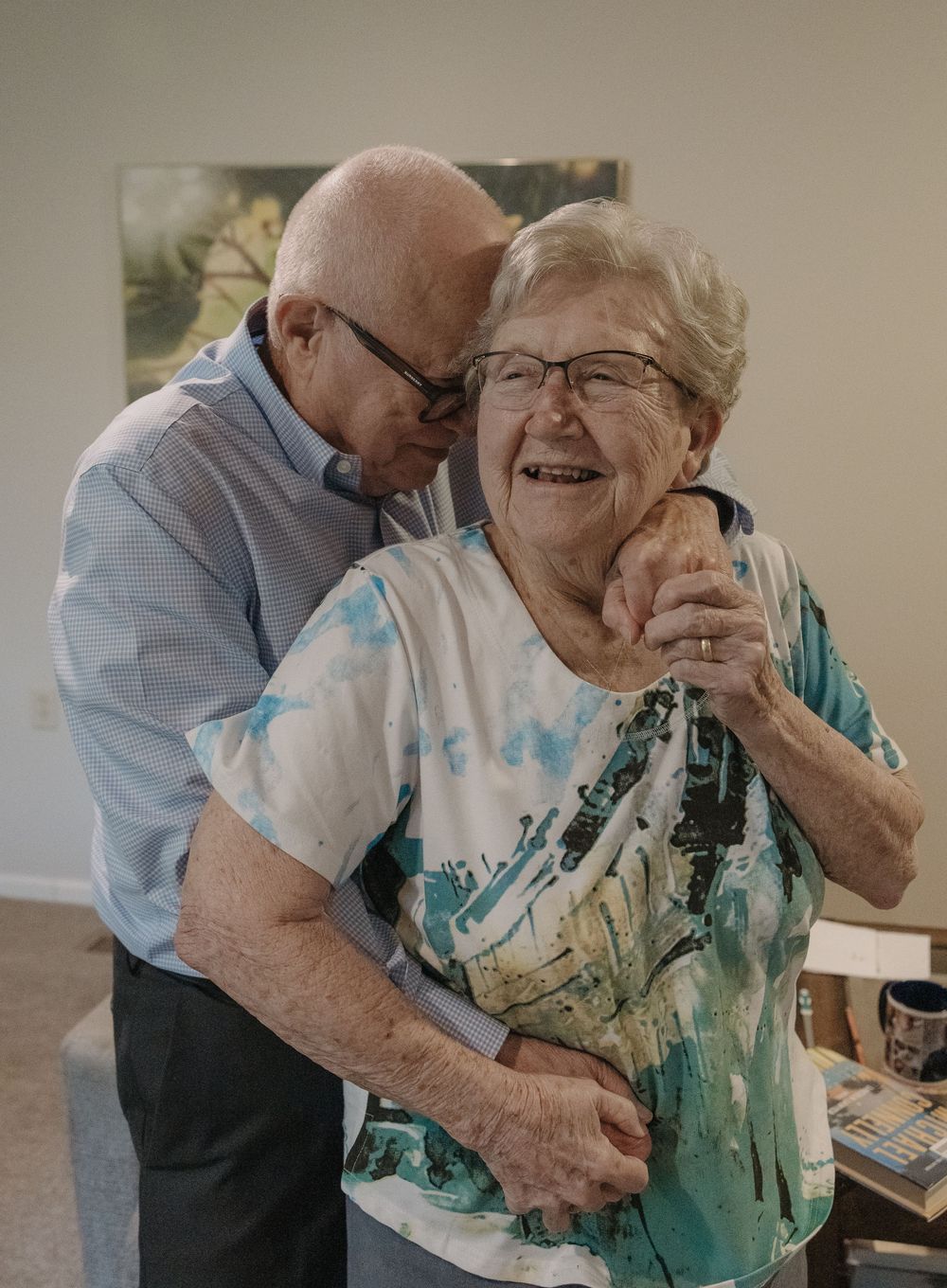 An elderly couple share a tender embrace while smiling warmly together, the person wearing a blue-green patterned blouse.